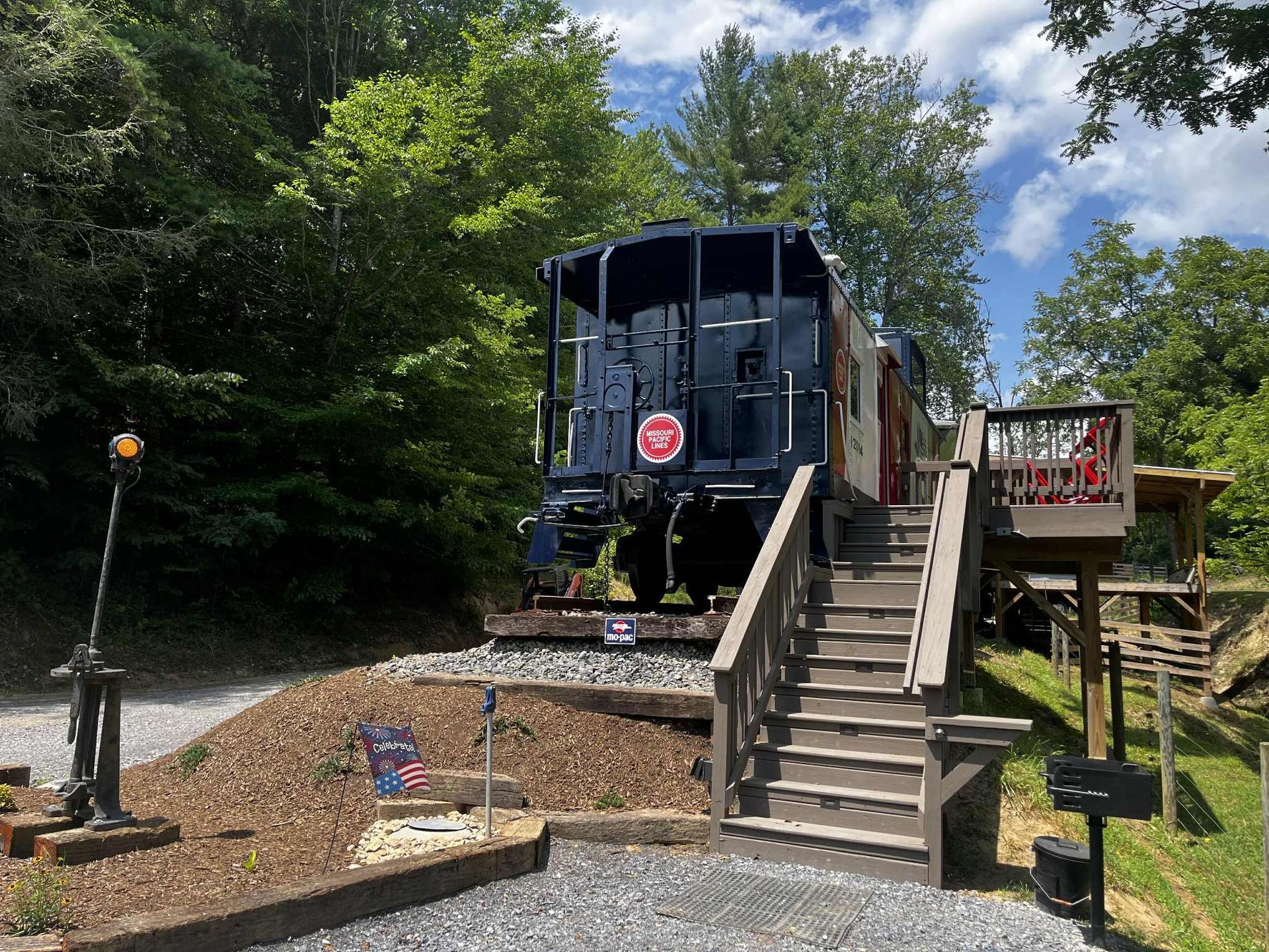 Blue train engine on tracks, wooden stairs lead up to a deck. Surrounded by trees.