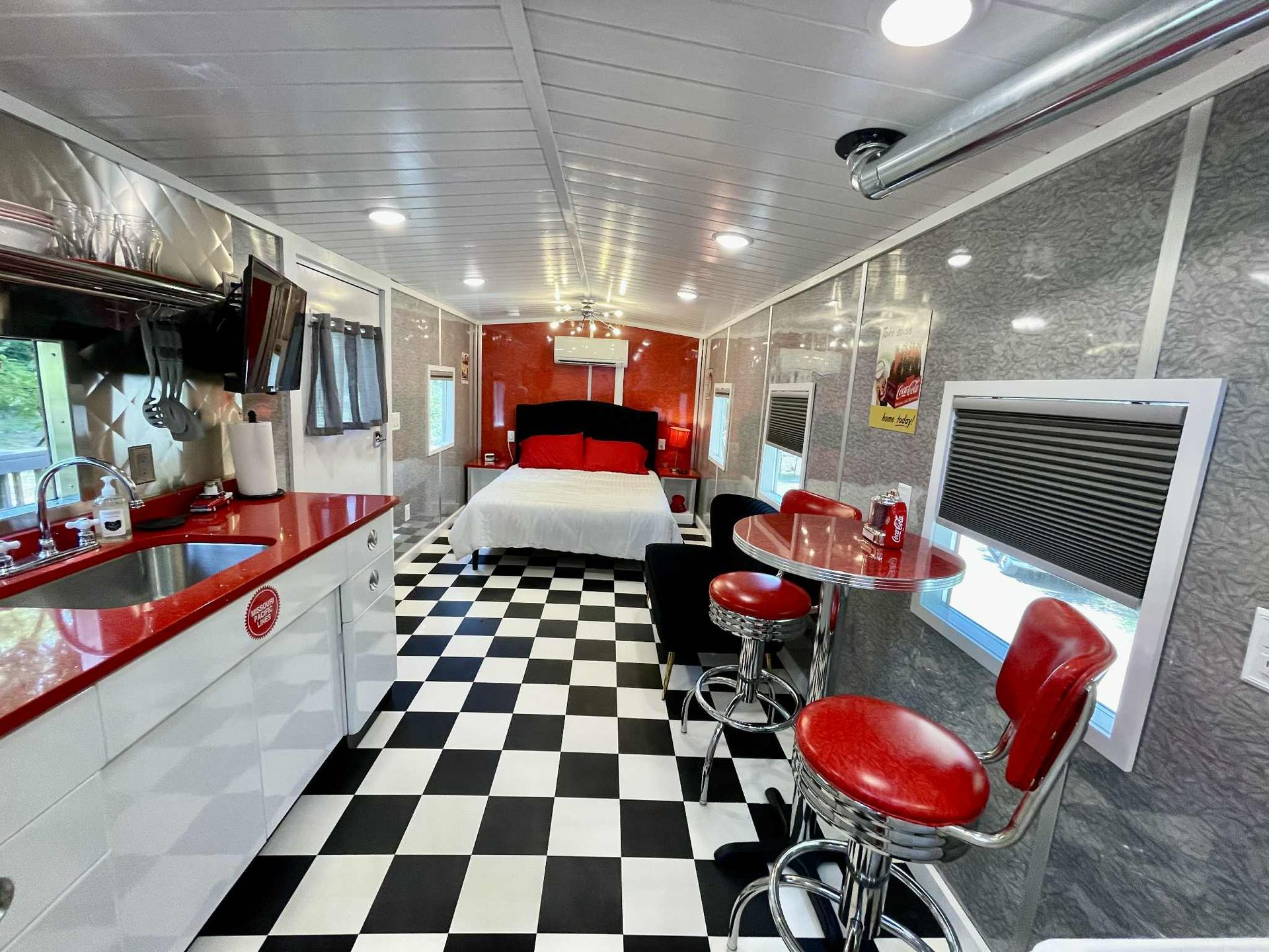 Retro-themed interior of a trailer: black and white checkered floor, red and silver accents, kitchen, bed, and dining set.