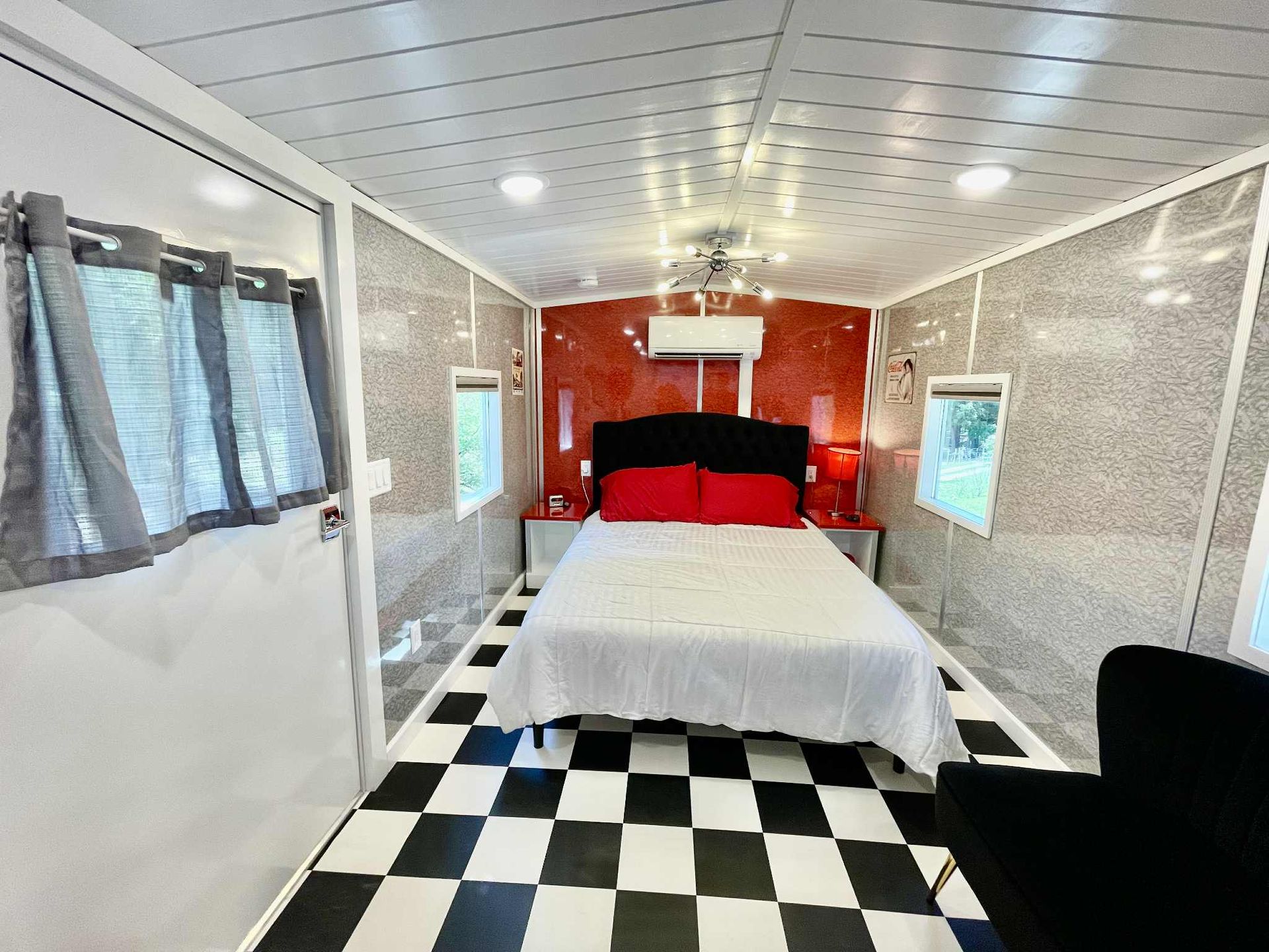 Bedroom in a converted trailer: black and white checkered floor, red accent wall, bed with red pillows.