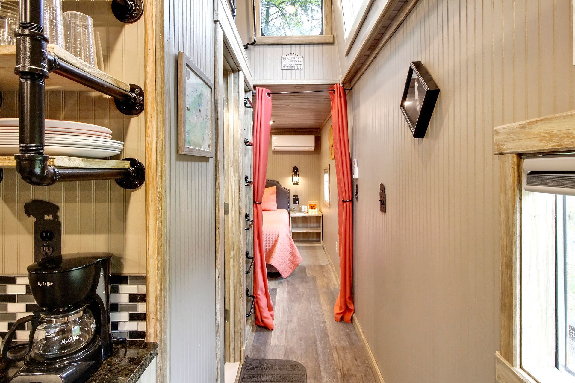 Narrow interior hallway of a tiny home with a bedroom at the end. Pink curtains and gray walls.