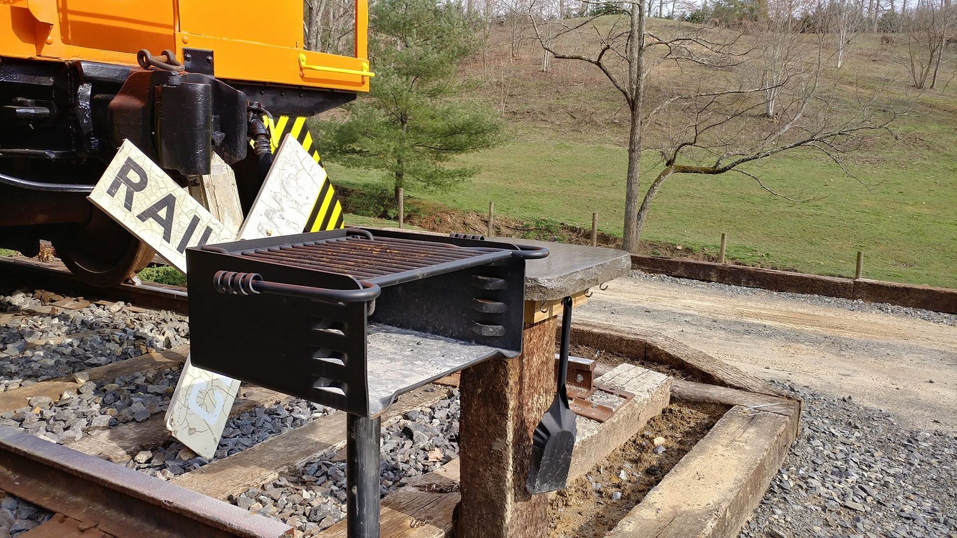 Black grill on a wooden stand next to railroad tracks and an orange train car.