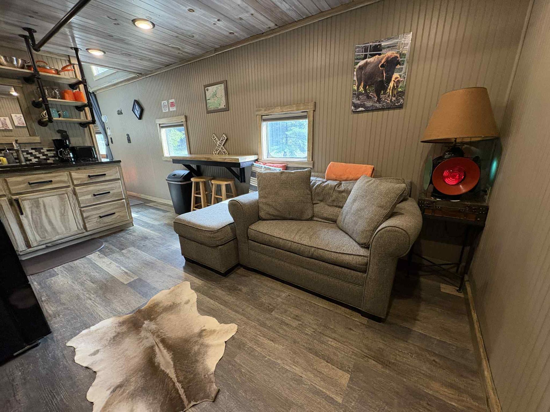 Cozy cabin interior: gray sofa, cowhide rug, small kitchen with shelving, small bar, and a lamp.