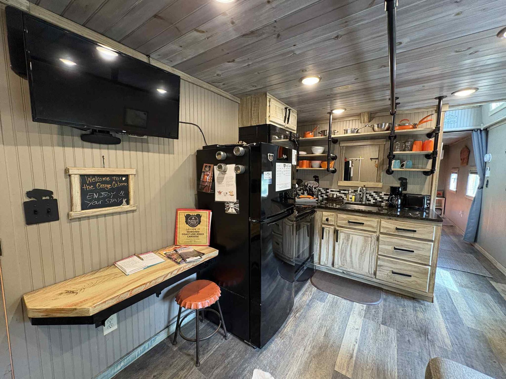 Interior of a tiny home with kitchen, TV, fold-down desk, and wooden accents.