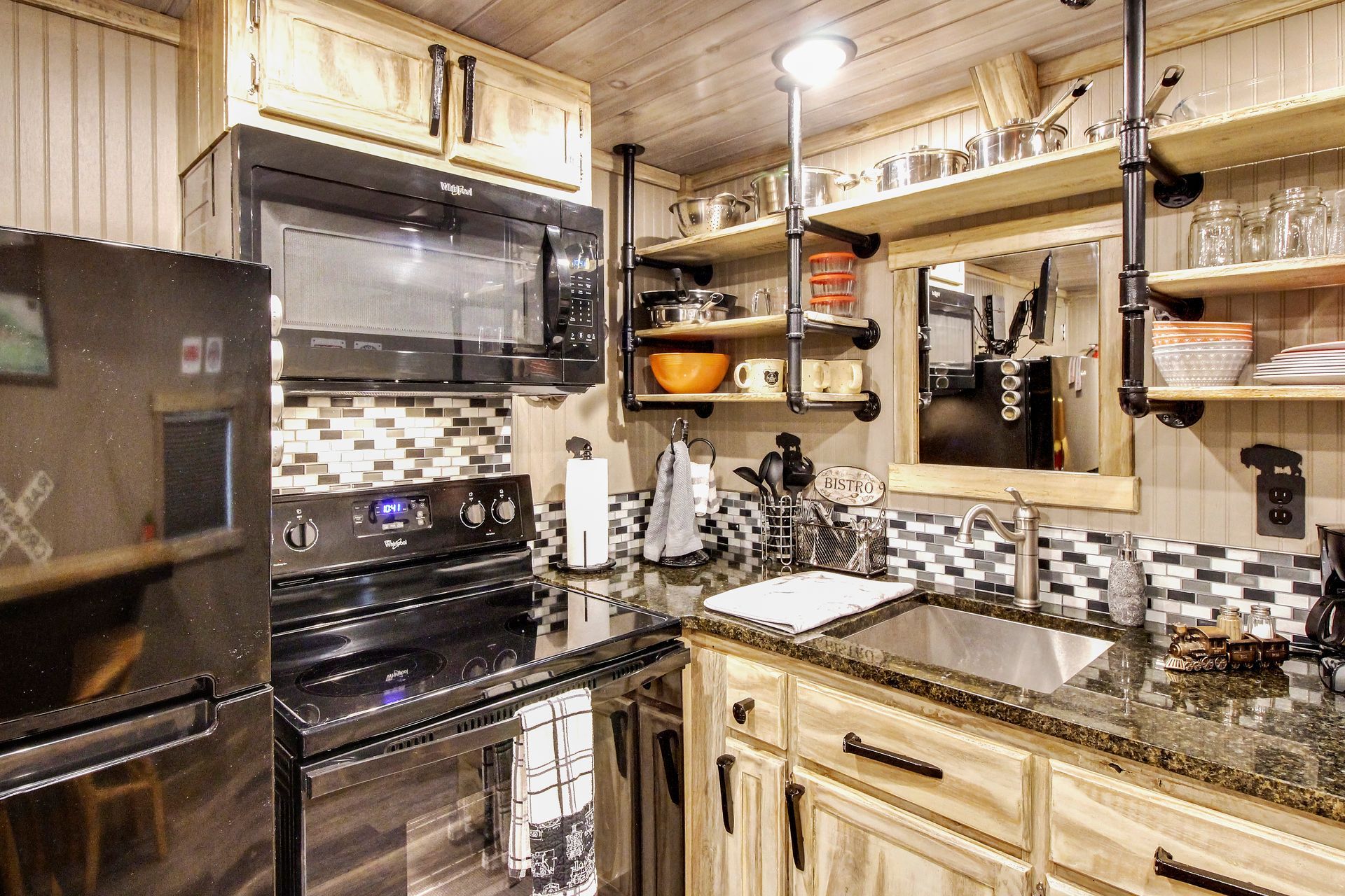 A small kitchen with wood cabinets, black appliances, and open shelving, plus a microwave, sink, and backsplash.