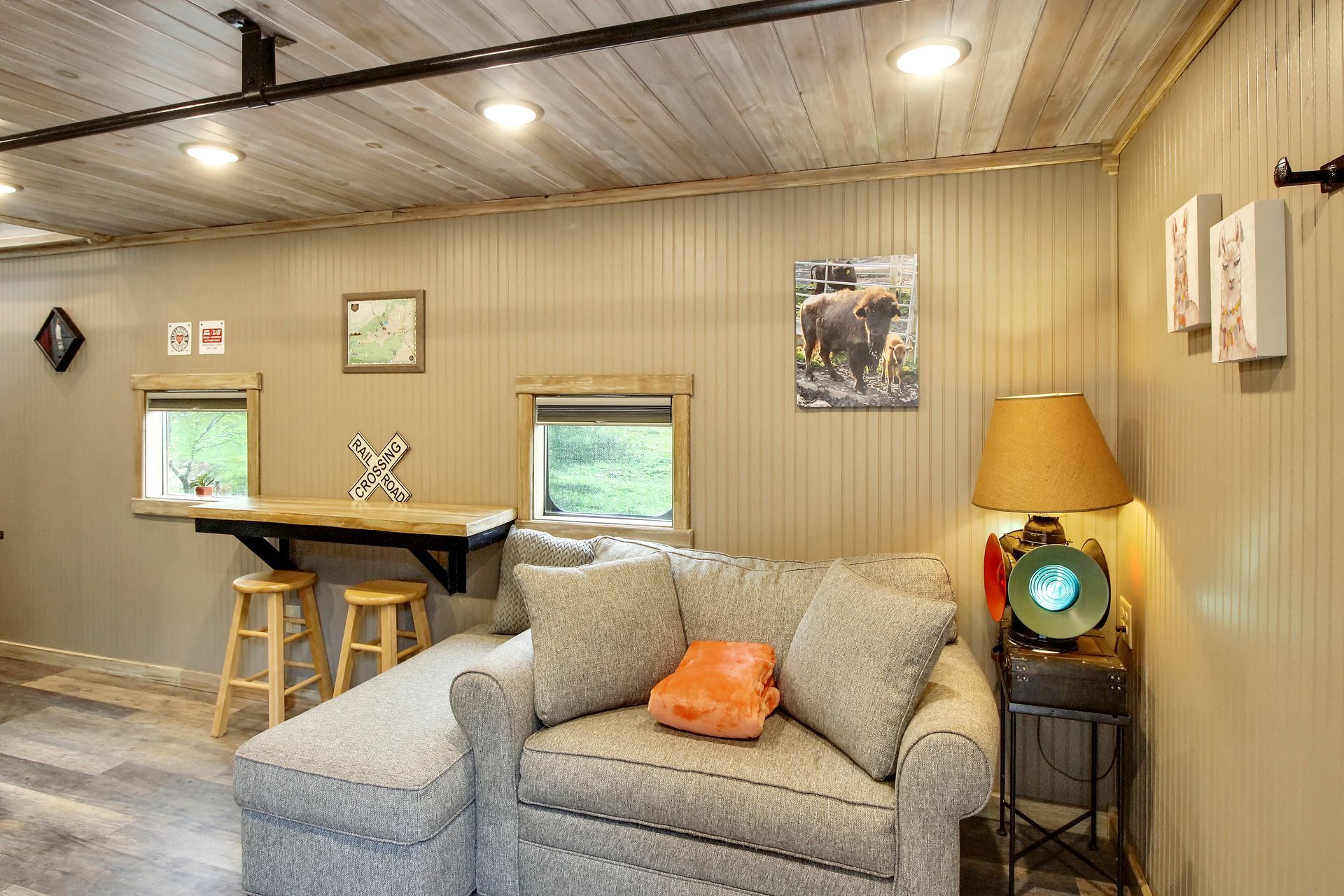 Cozy living room with gray couch, small windows, bar, wooden ceiling, and a lamp.