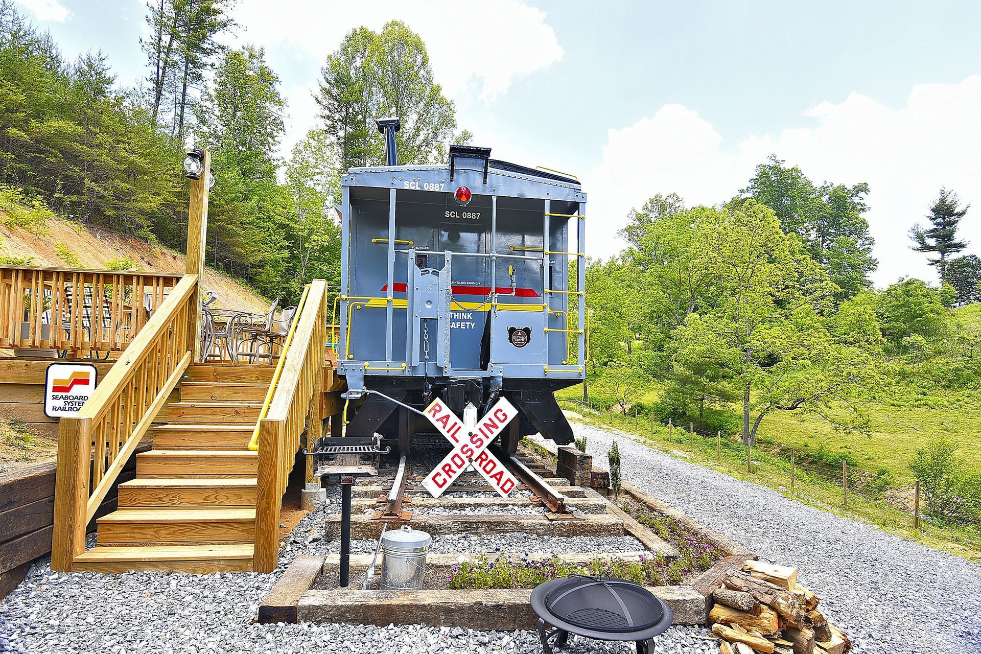 Grey caboose on tracks with wooden stairs, crossbuck sign, and fire pit in a natural setting.
