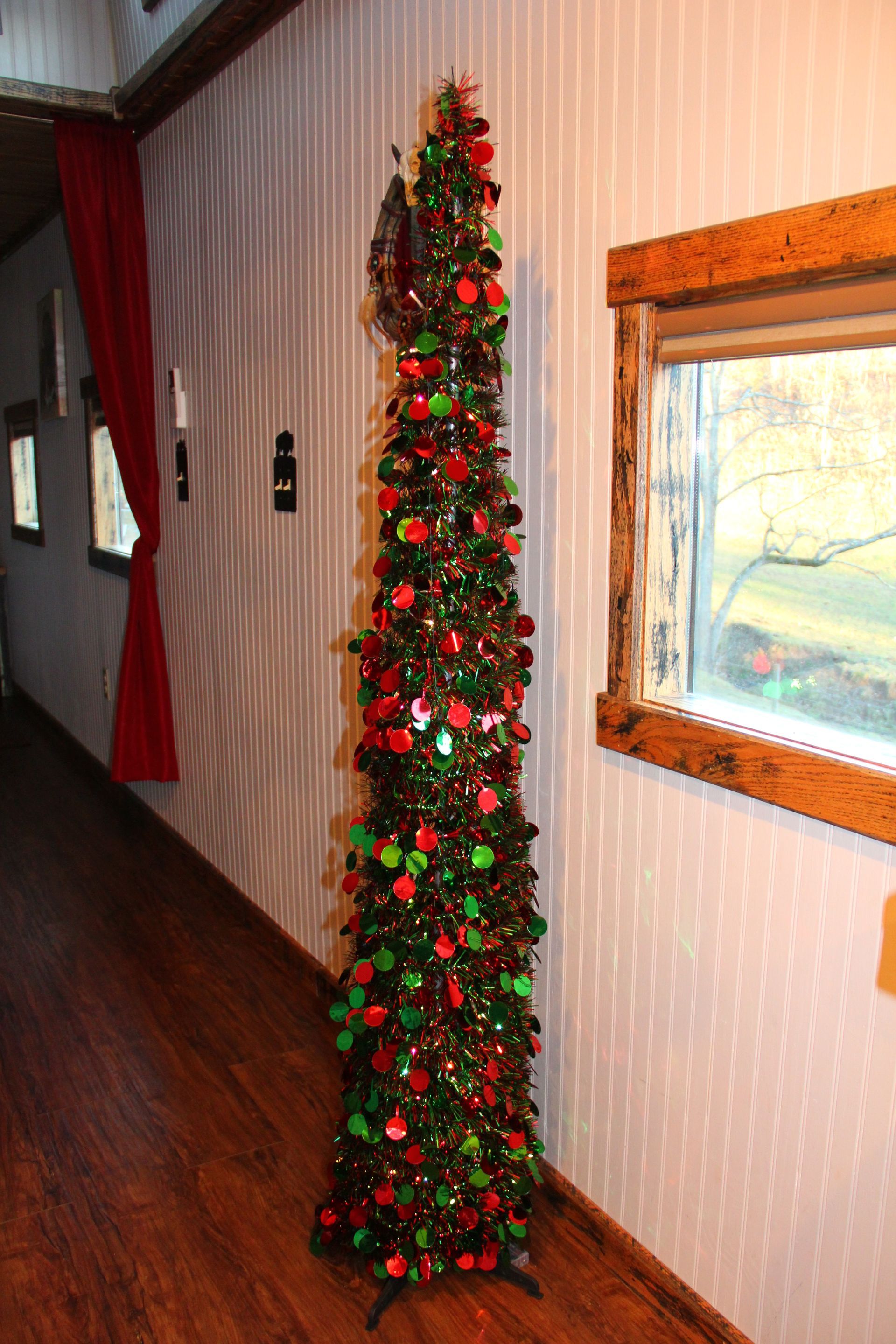 Tall, slim Christmas tree decorated with red, green, and gold ornaments, in a hallway with a red curtain and window.