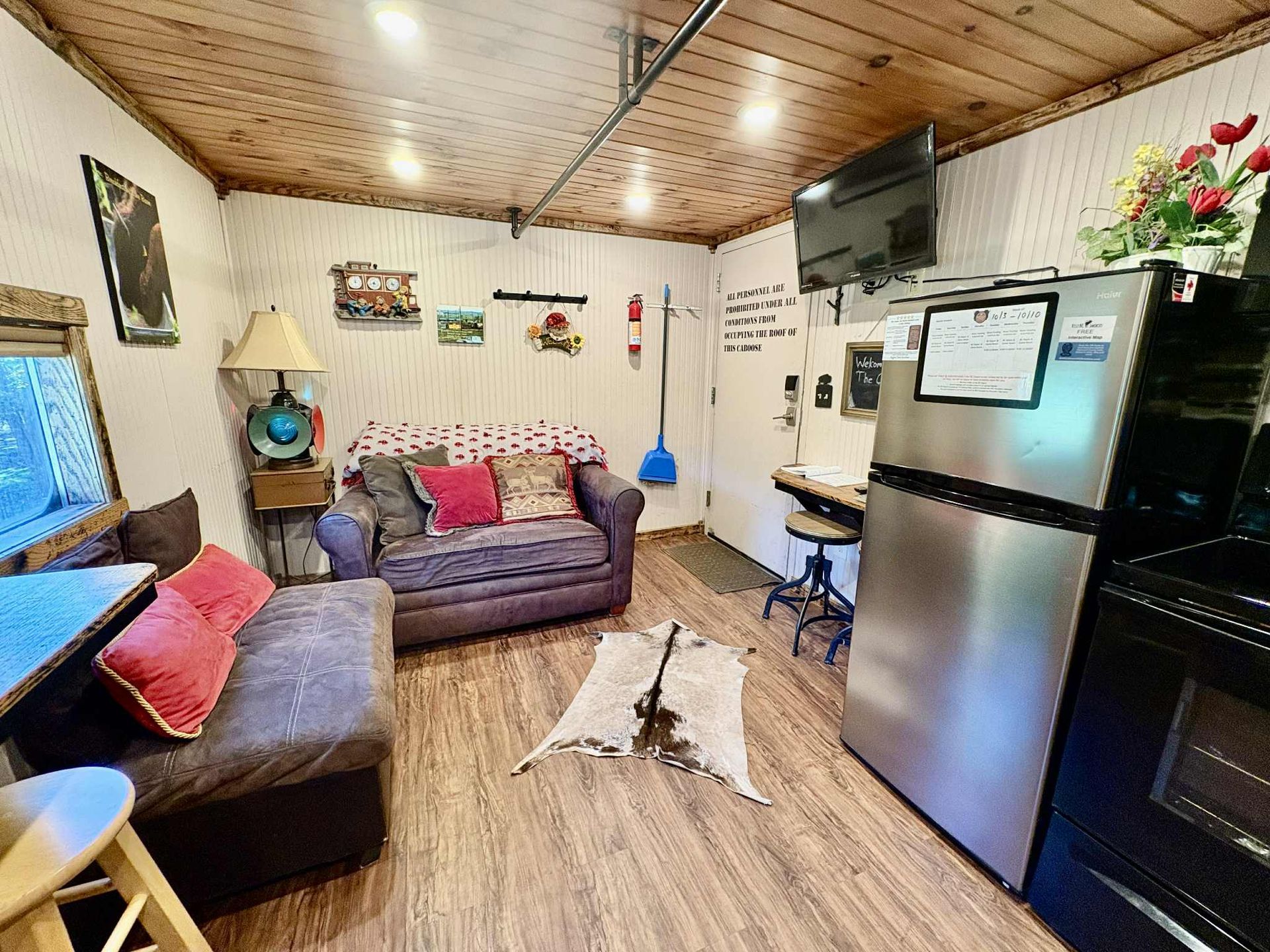 Cozy cabin interior: sofa, desk, fridge, wooden ceiling, rug, decor.