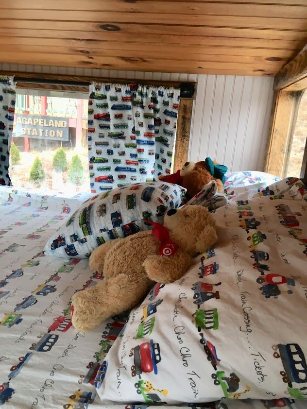 Teddy bears and pillows on a bed with a train-themed sheet set. Wooden ceiling and window.