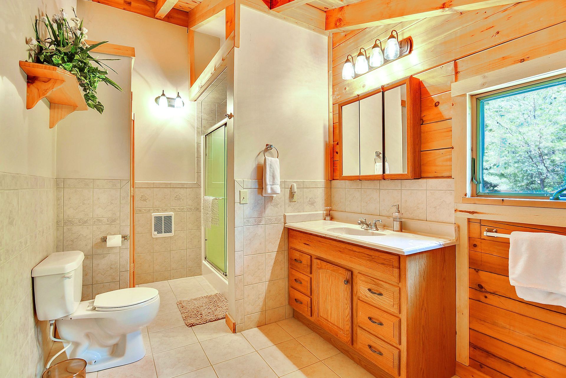 Bathroom with wooden walls and cabinets, featuring a toilet, sink, shower, and a small window with a towel rack.
