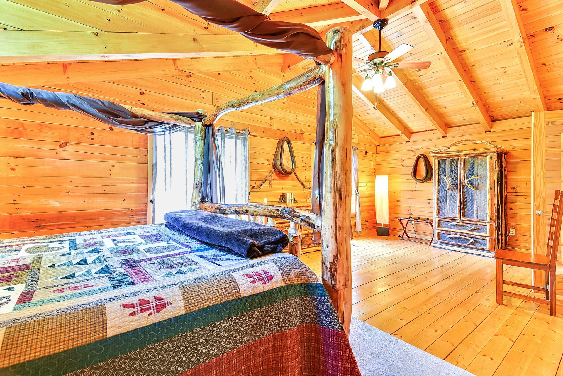 Bedroom with log bed frame, wood walls, and ceiling. A quilt, two windows, a chair and a ceiling fan are visible.
