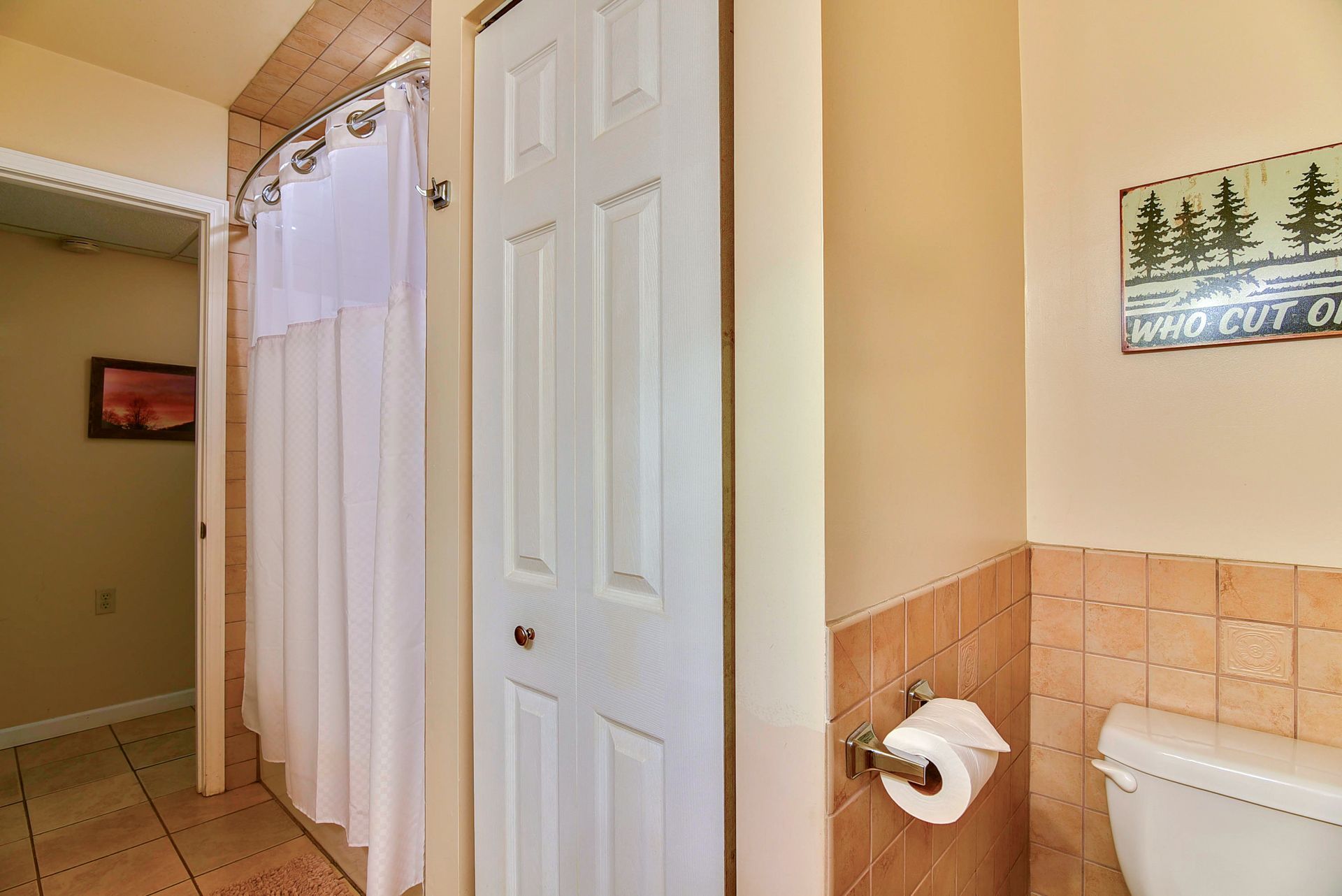 Bathroom with a closed white door, shower with white curtain, toilet, and a sign depicting trees.
