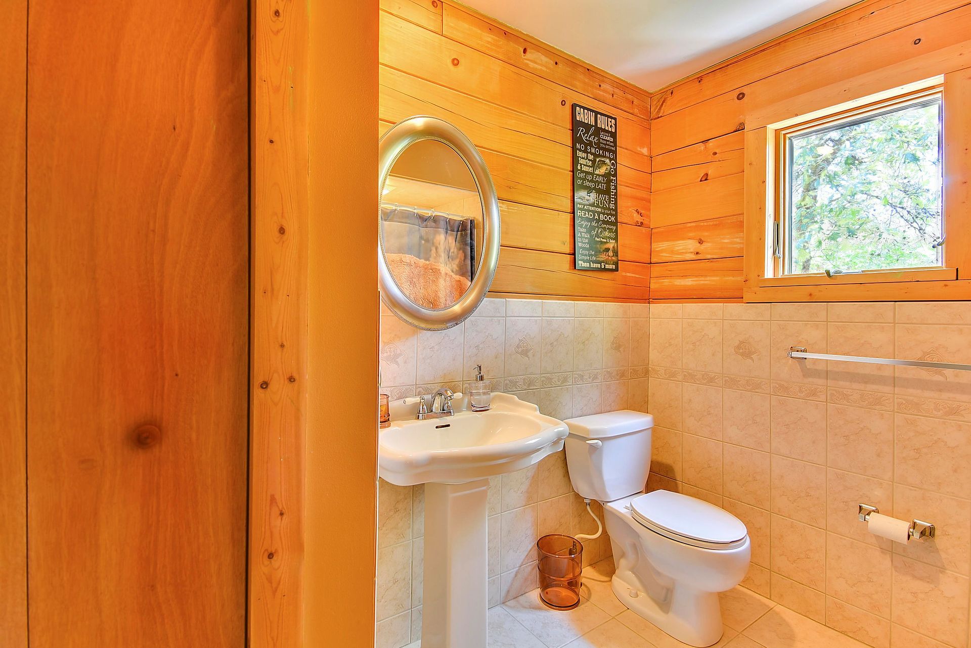 Bathroom with wood paneling, pedestal sink, toilet, oval mirror, and small window.