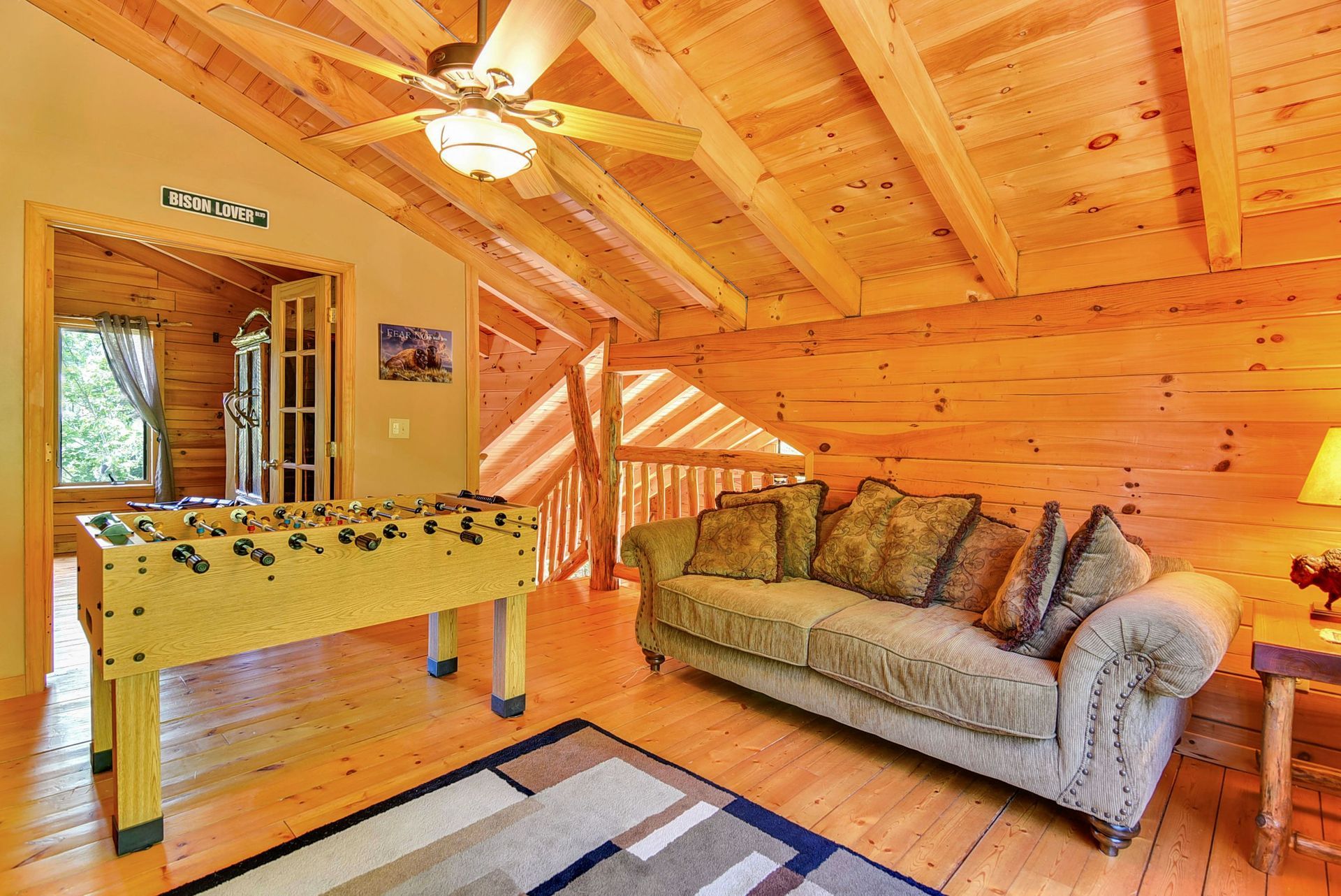 Cozy cabin loft with foosball table, sofa, and wooden walls/ceiling. Sunlight streams in.