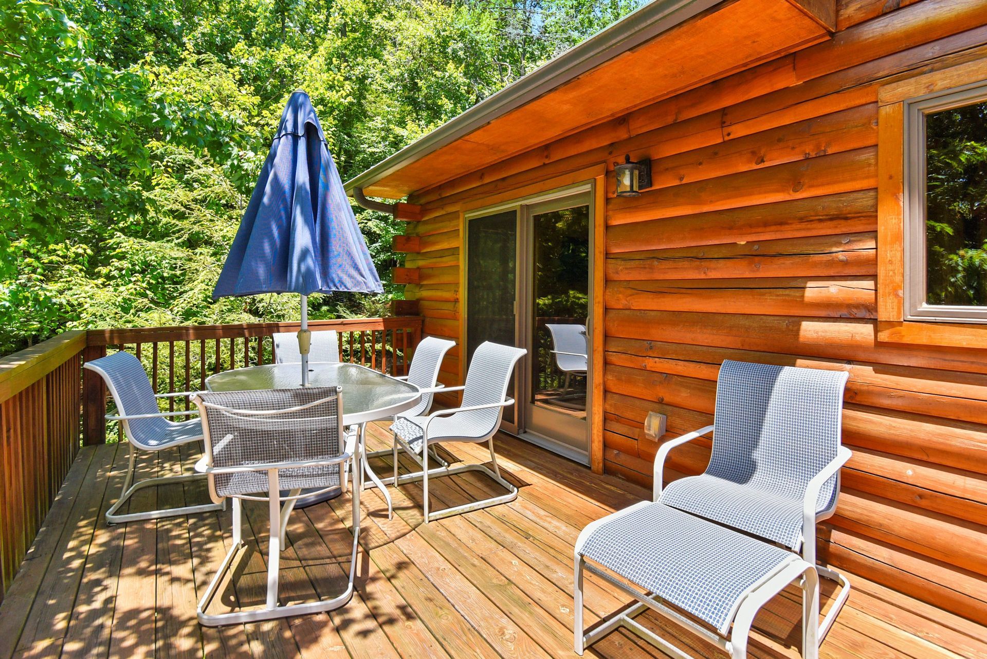 Wooden deck with outdoor furniture, blue umbrella, next to a cabin with sliding glass door.