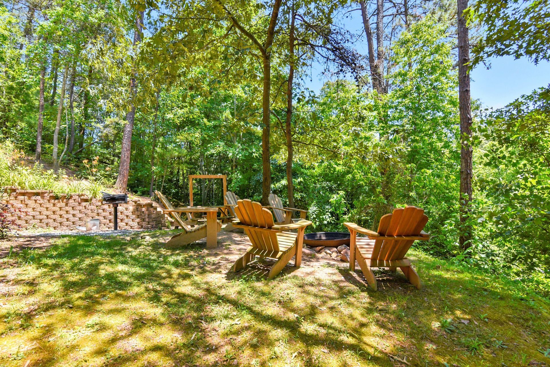 Two wooden Adirondack chairs face a small fire pit, surrounded by trees and greenery.