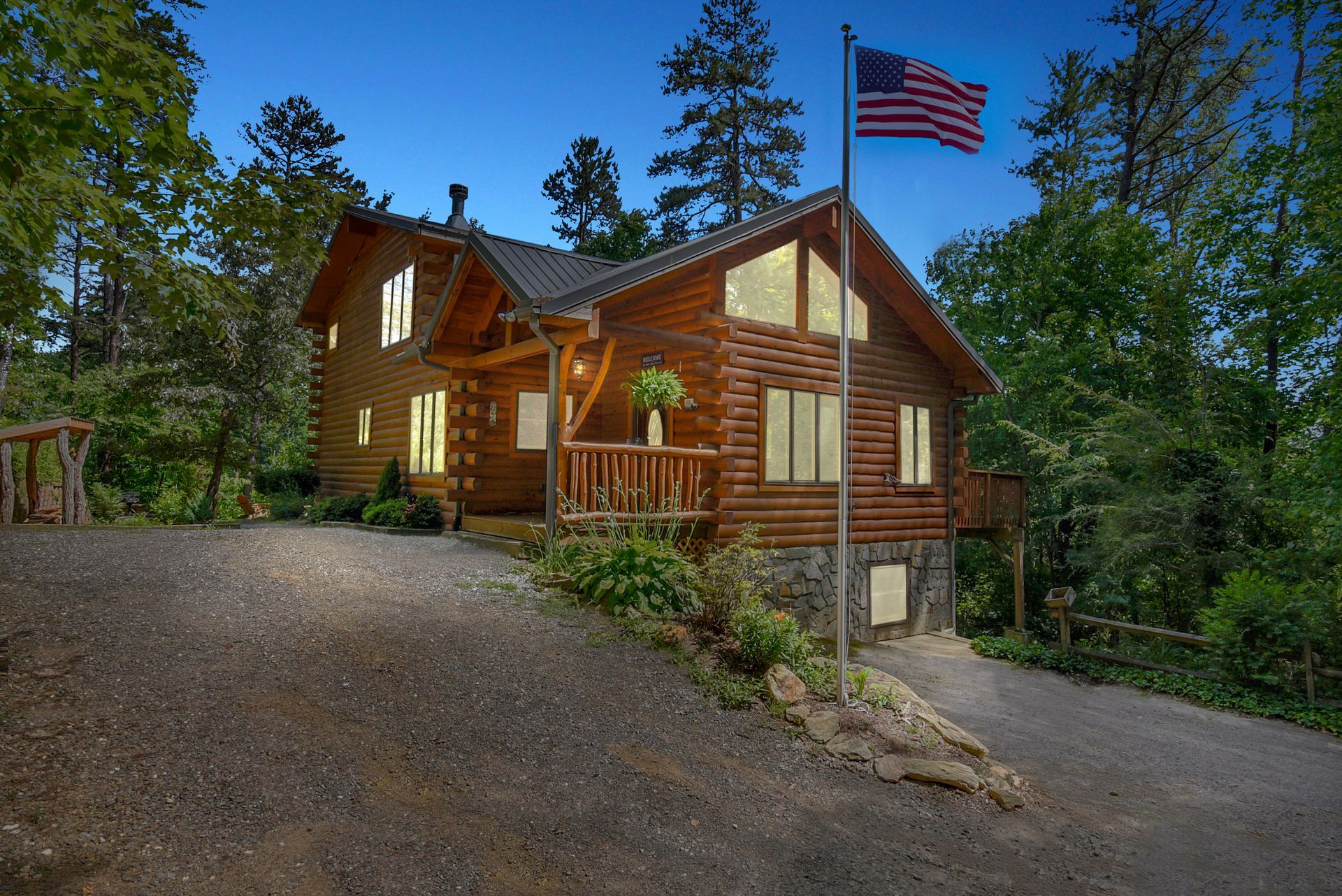 Log cabin with American flag, driveway, and surrounding trees. Dusk setting.