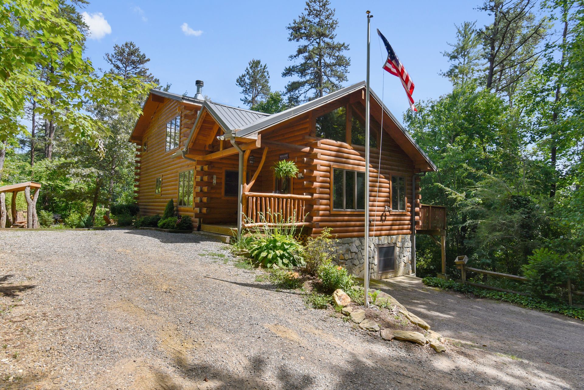 Log cabin with American flag, surrounded by trees, gravel driveway.