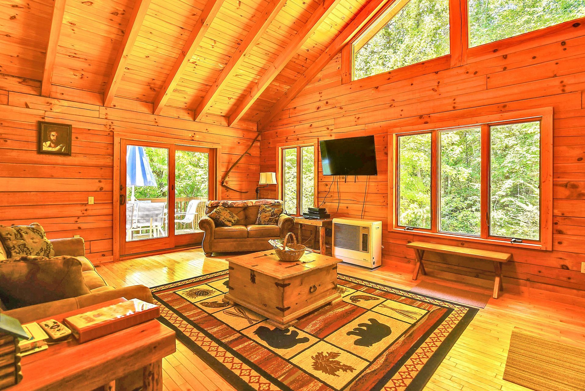 Cozy cabin living room with wooden walls, a rug, a sofa, and windows overlooking greenery.