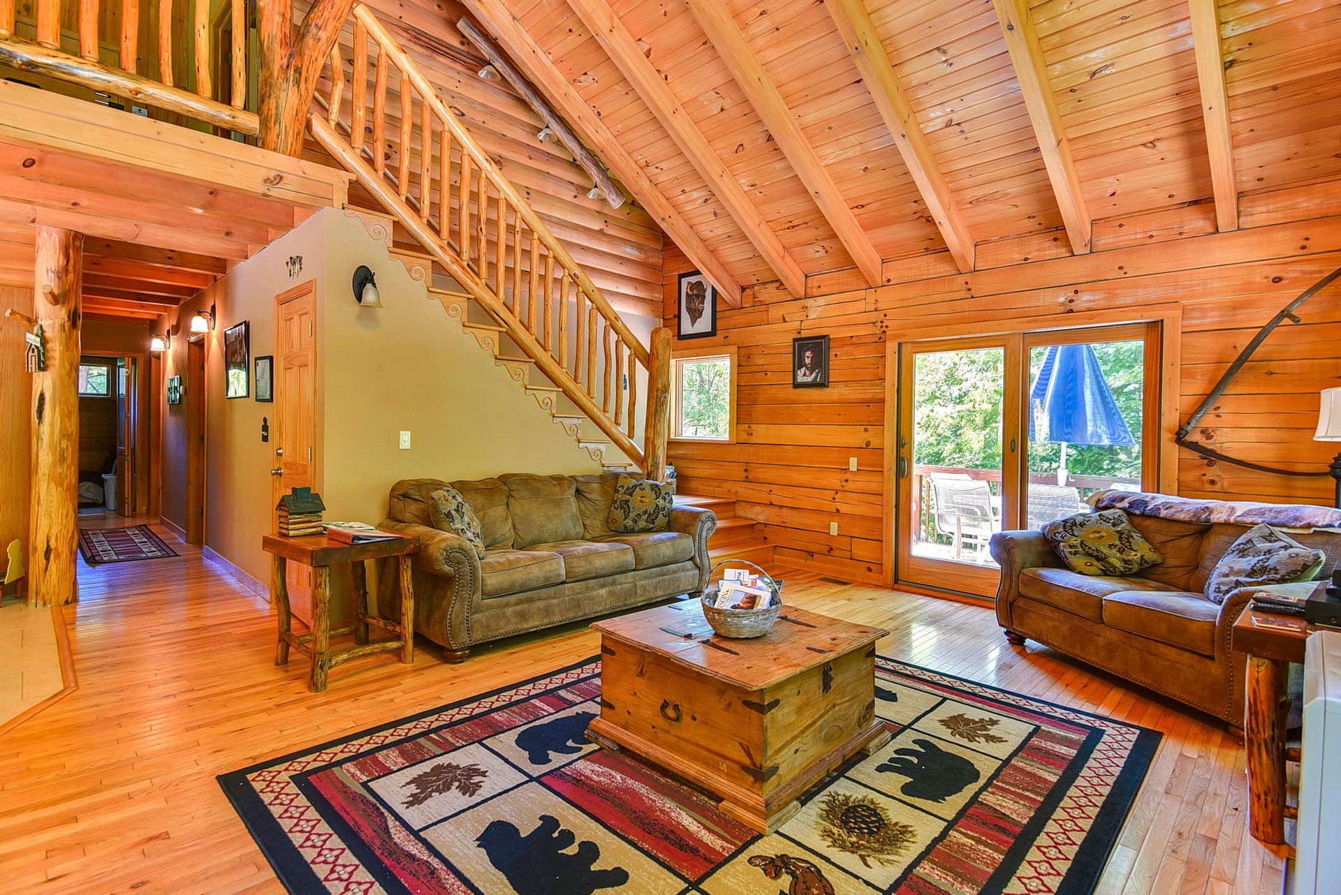 Cozy wooden cabin interior with a sofa, rug, staircase, and a doorway to an outdoor space.