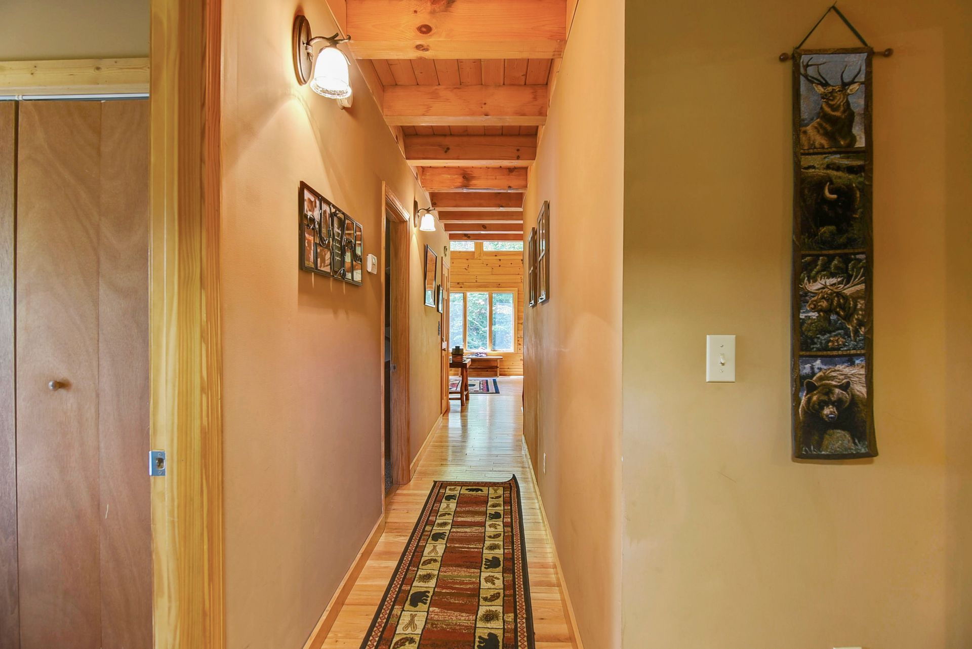 Narrow hallway with wood ceiling, beige walls, and a rug. Light fixtures and framed artwork line the walls.