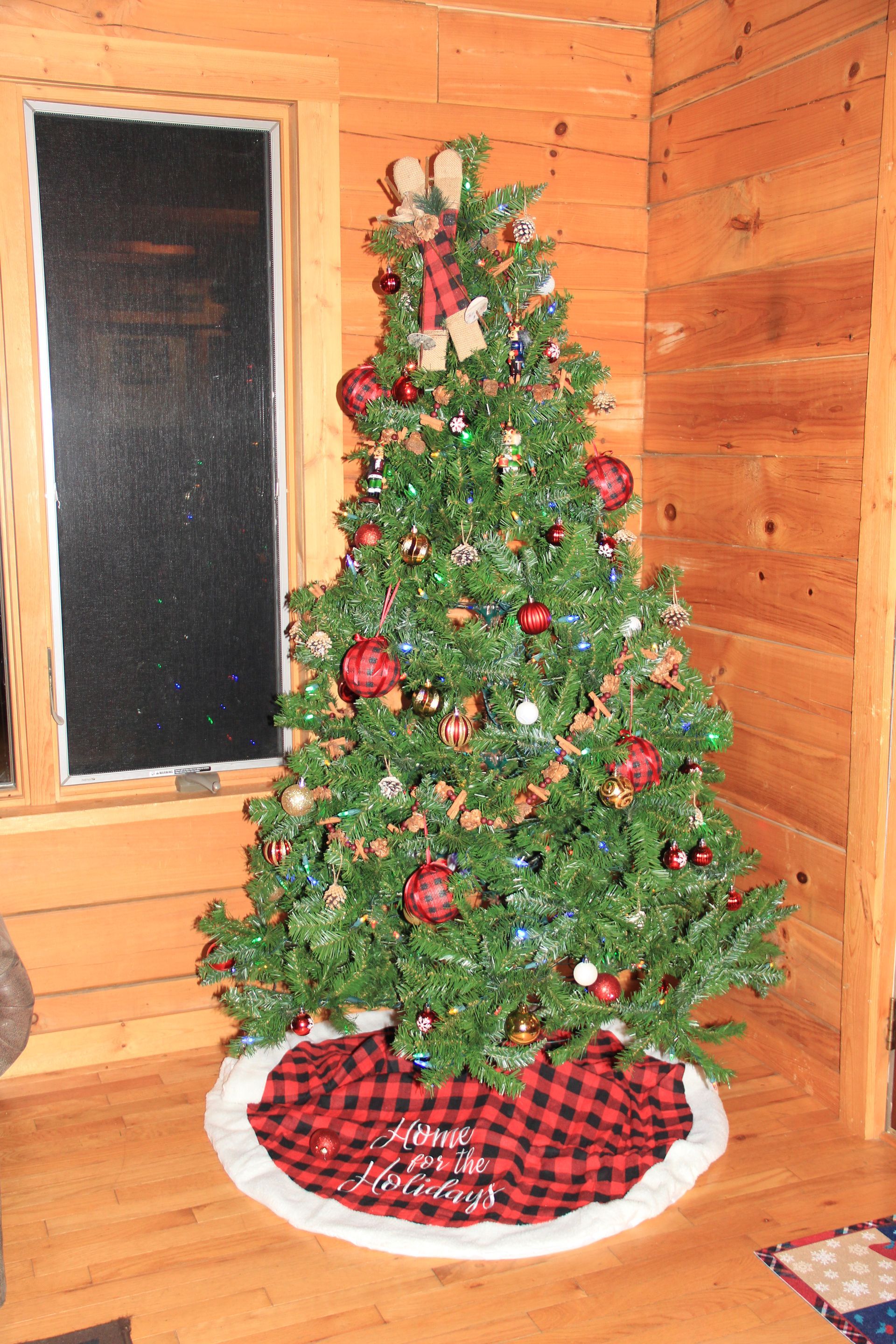 Christmas tree decorated with red ornaments and plaid skirt, set in a log cabin.