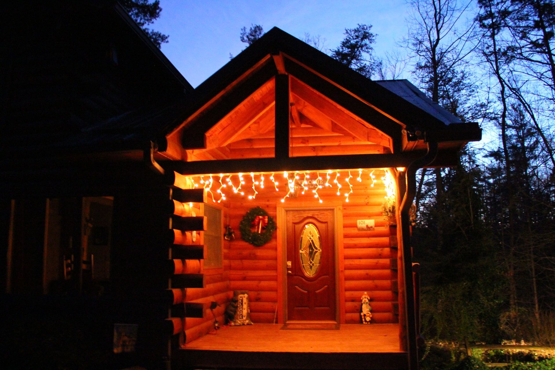 Log cabin porch lit with string lights and a wreath on the door at dusk.