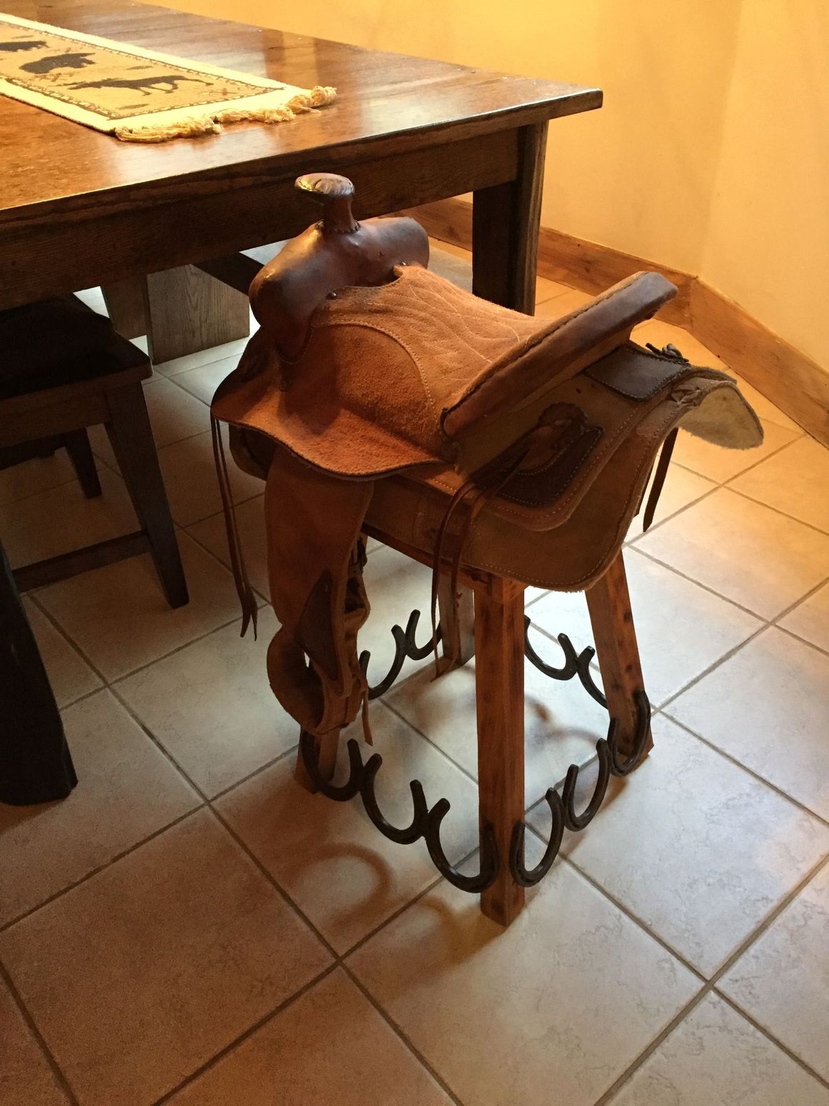A western saddle used as a stool next to a wooden table with tile flooring.