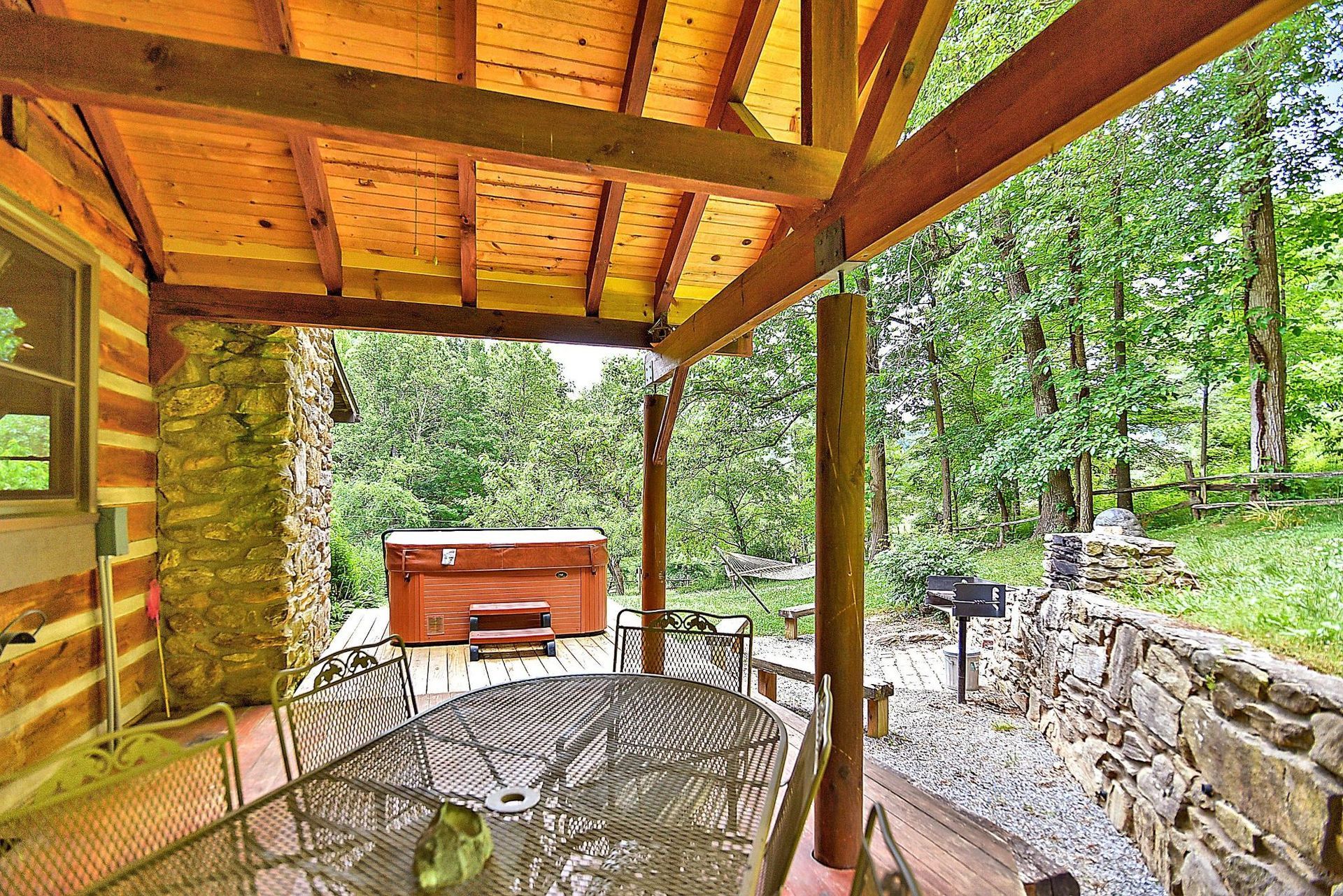 Covered porch with hot tub and grill, surrounded by trees and stone walls.