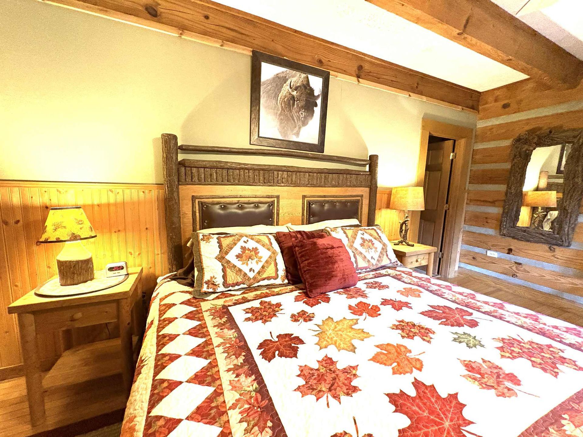 Bedroom with a rustic bed, autumn-themed quilt, log walls, framed bison art, and nightstand lamps.