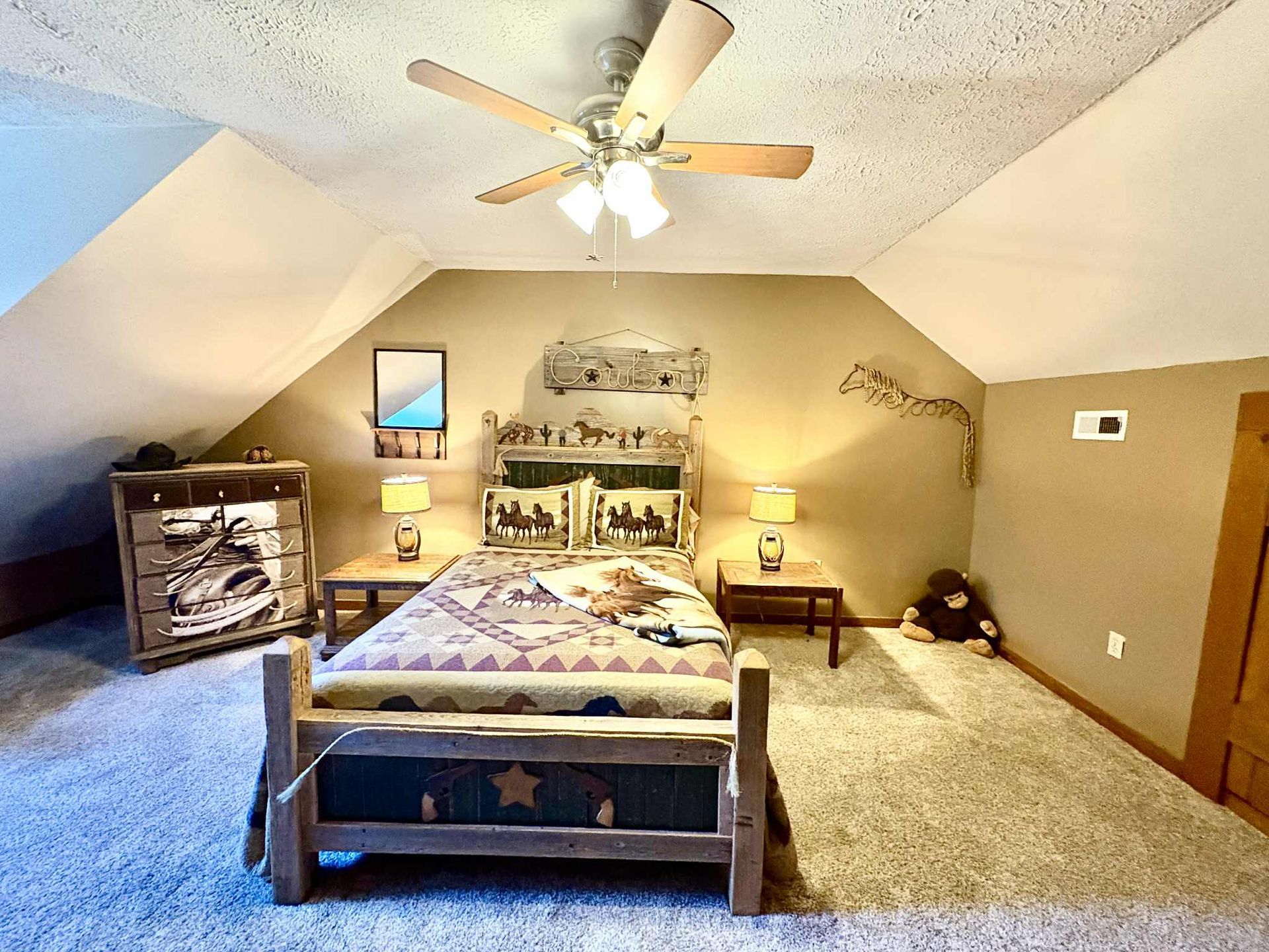 Bedroom with bed, dresser, and ceiling fan under a slanted roof. Earthy tones and rustic furniture.