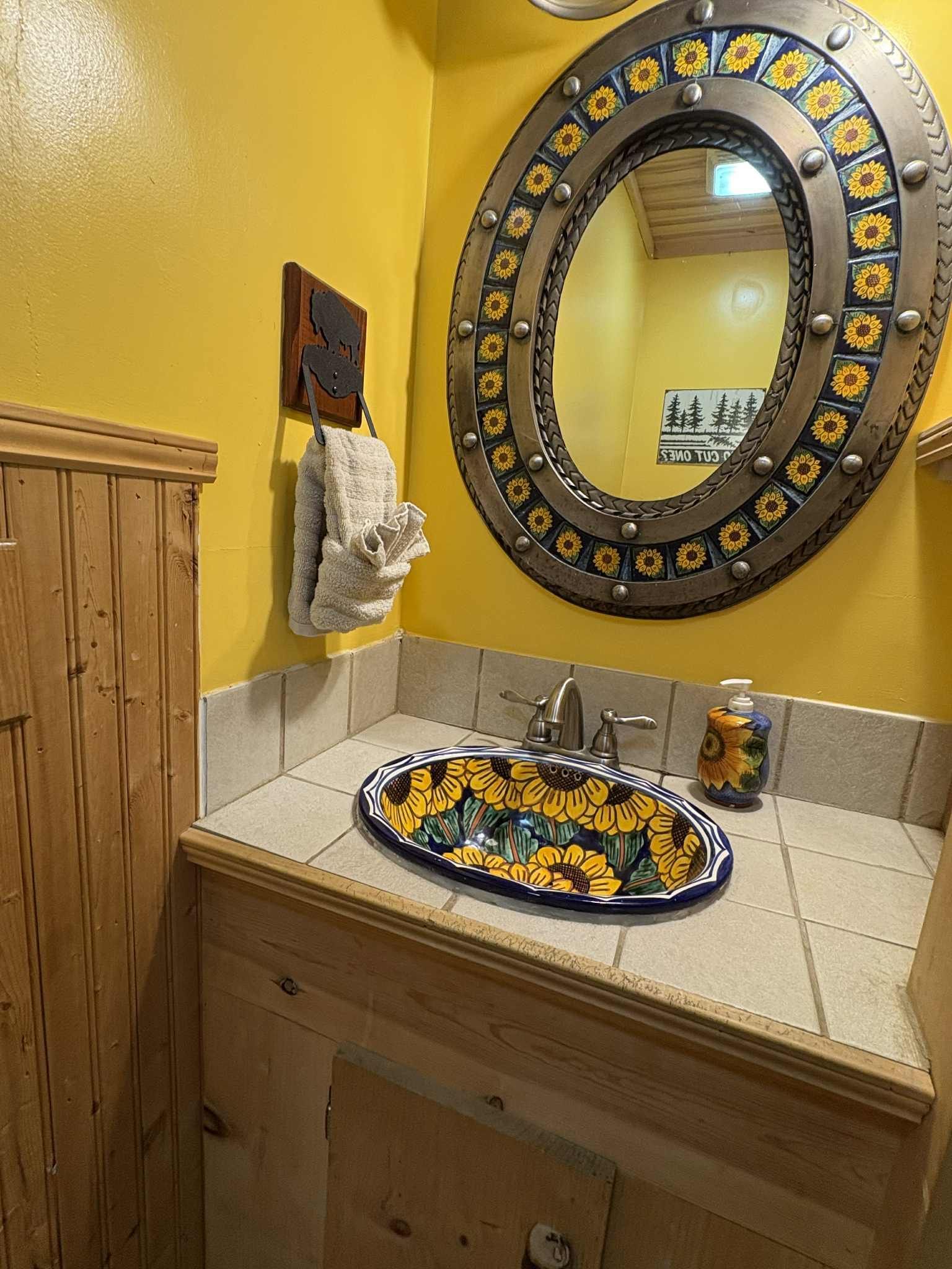 A bathroom with a yellow wall, a sunflower sink, a decorative mirror, and a wooden cabinet.
