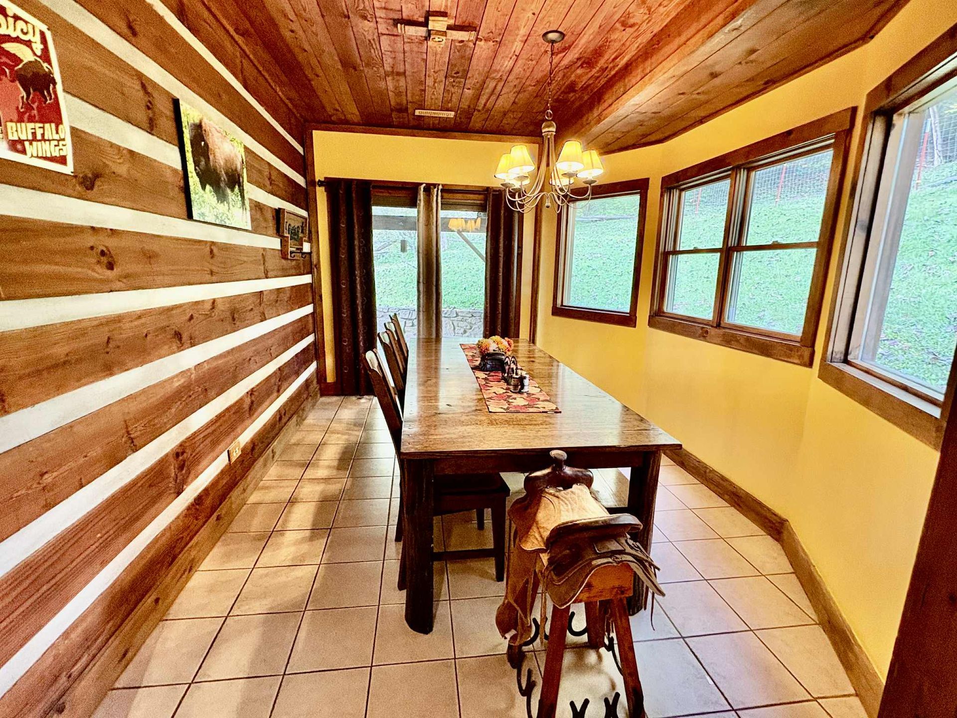 Dining room with log walls, wood ceiling, table, saddle, and windows overlooking greenery.