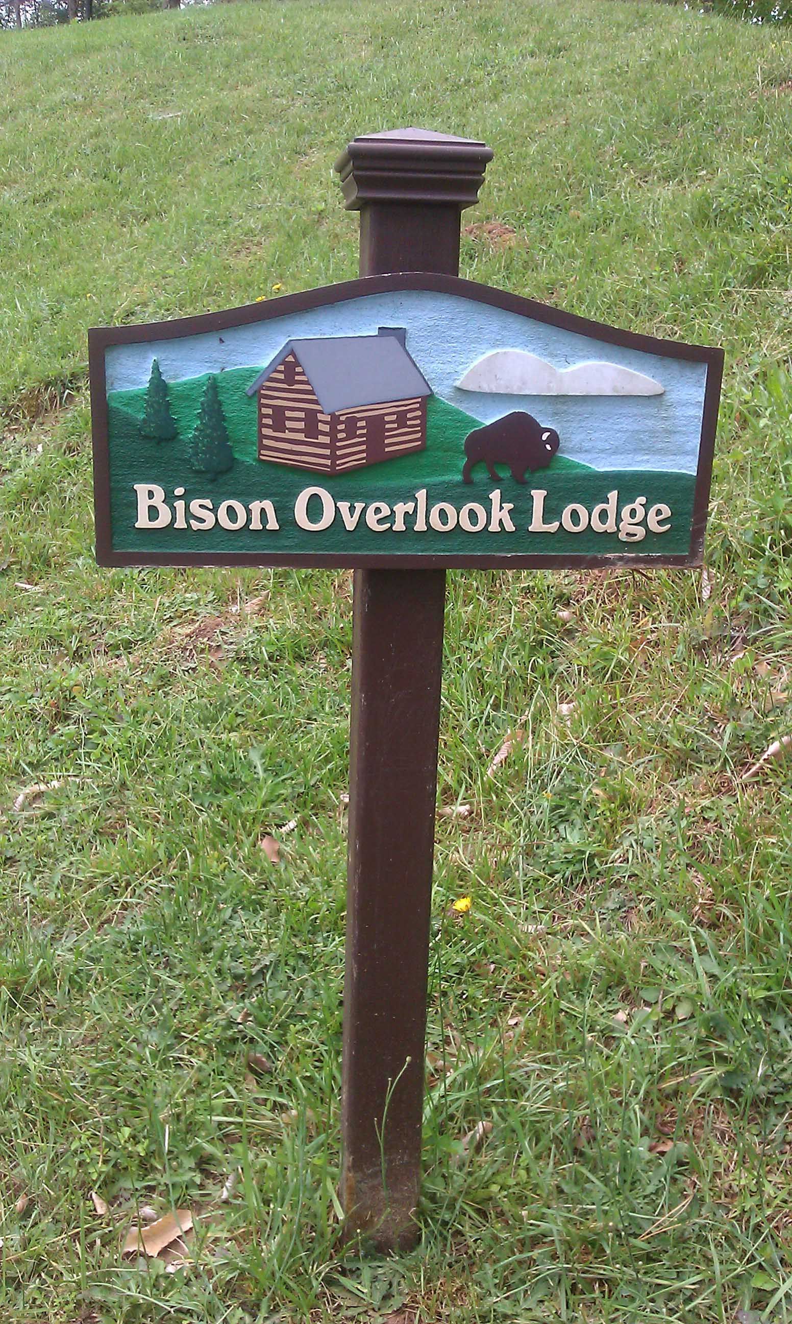 Sign for Bison Overlook Lodge: brown post, painted sign with log cabin, bison, trees, sky; text