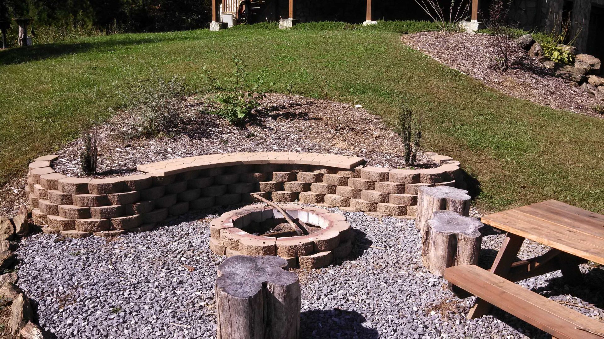 Fire pit area with stone bench and seating, surrounded by gravel, grass, and a wooden picnic table.