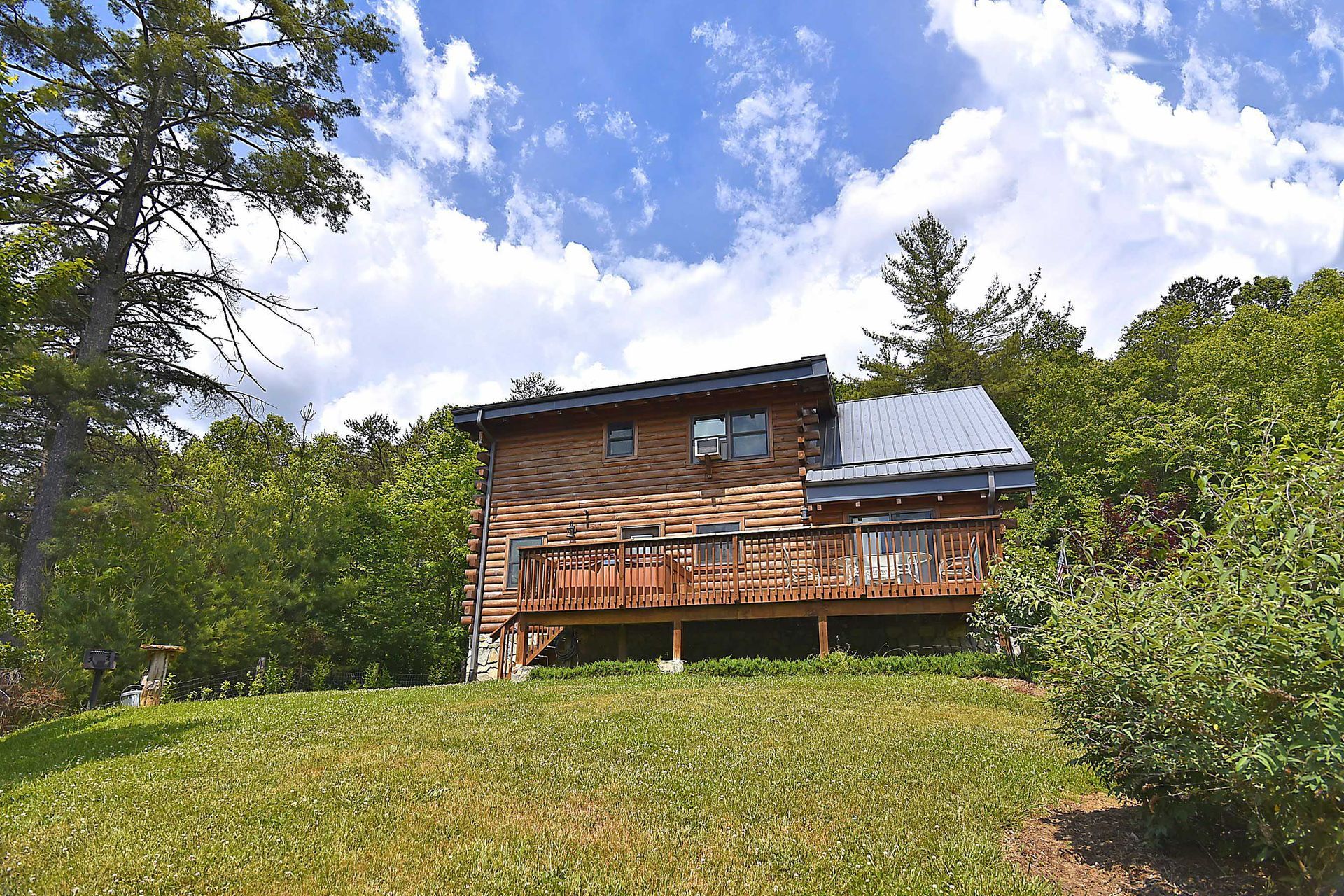 Cabin with wooden deck, surrounded by trees, on a grassy hill under a blue sky.