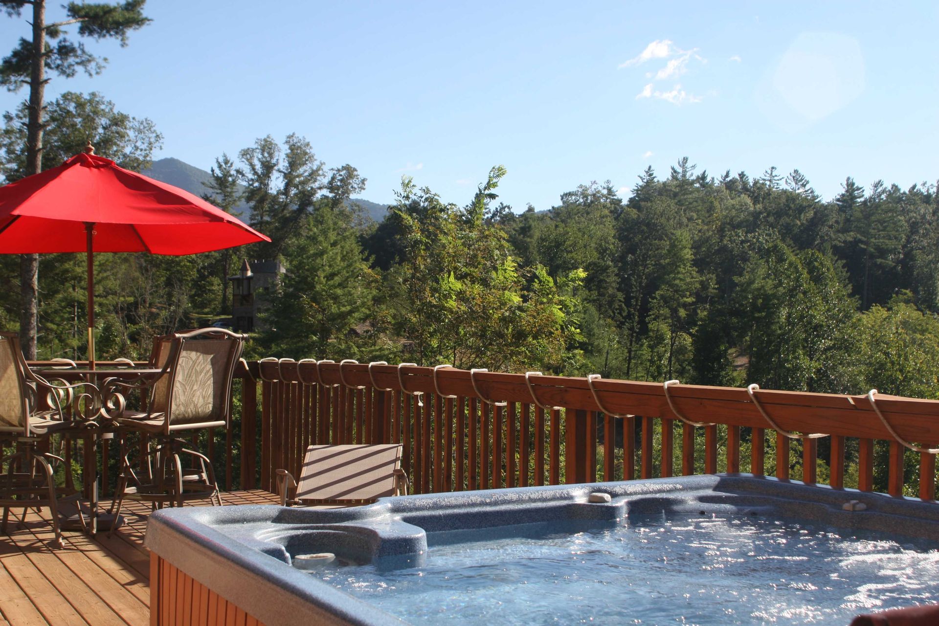 Hot tub on a wooden deck with a red umbrella, overlooking a green forest on a sunny day.
