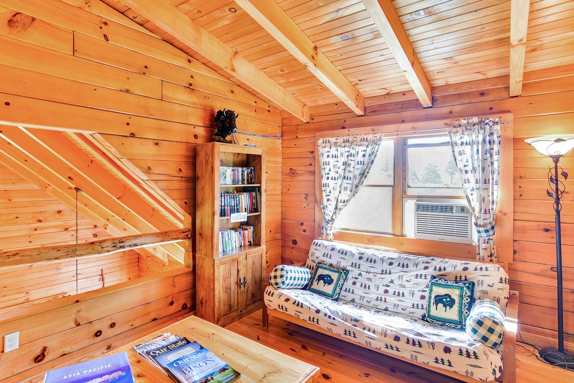 Cozy cabin interior with wooden walls, futon, bookshelf, and a window with curtains.