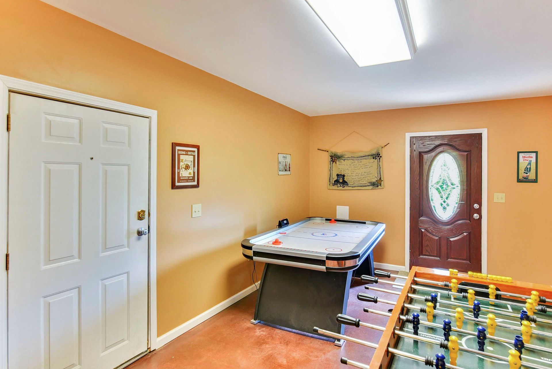 Game room with an air hockey table, foosball, and two doors against orange walls.