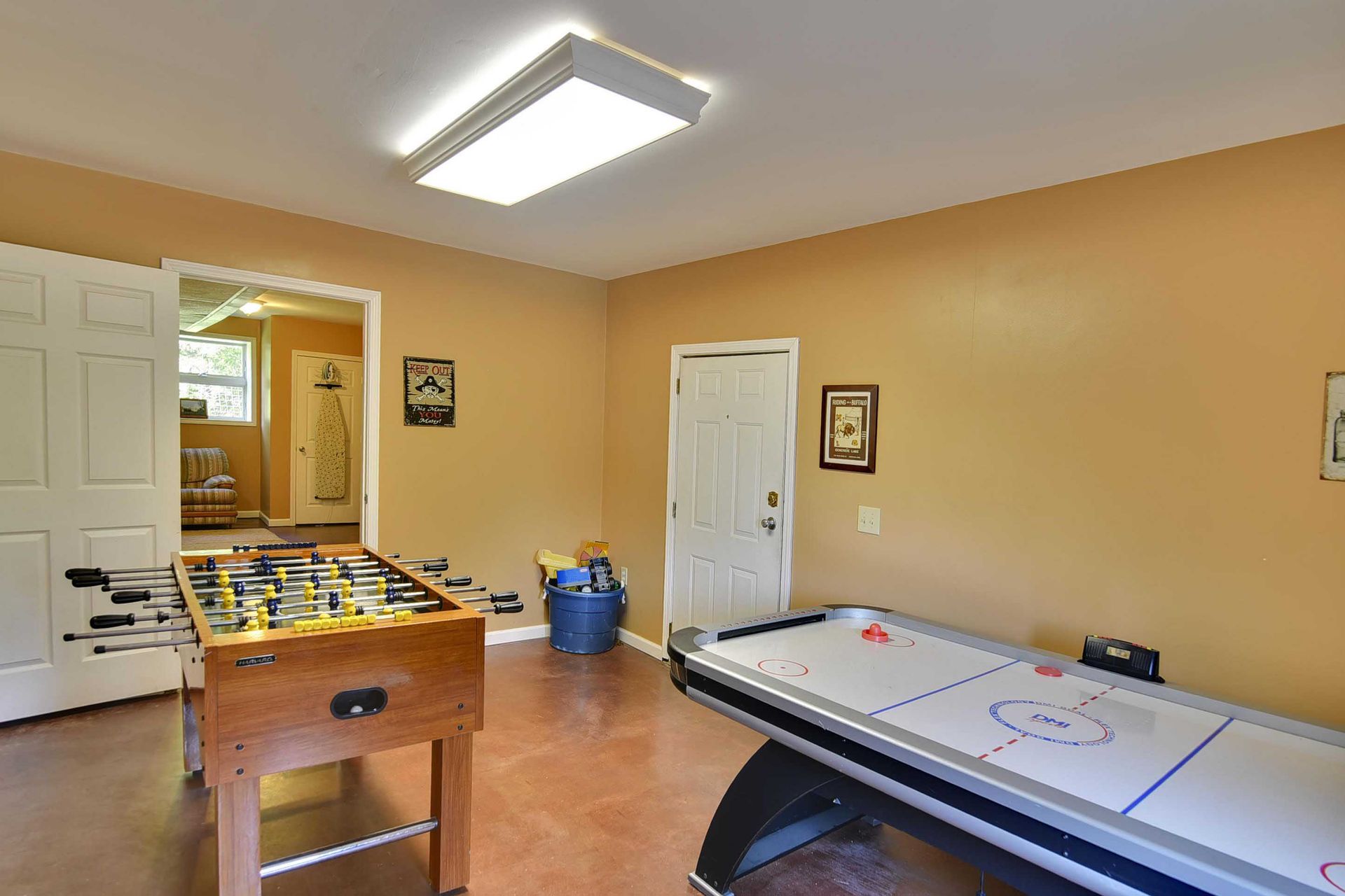 Game room with foosball and air hockey tables; tan walls, white doors, and a bright overhead light.
