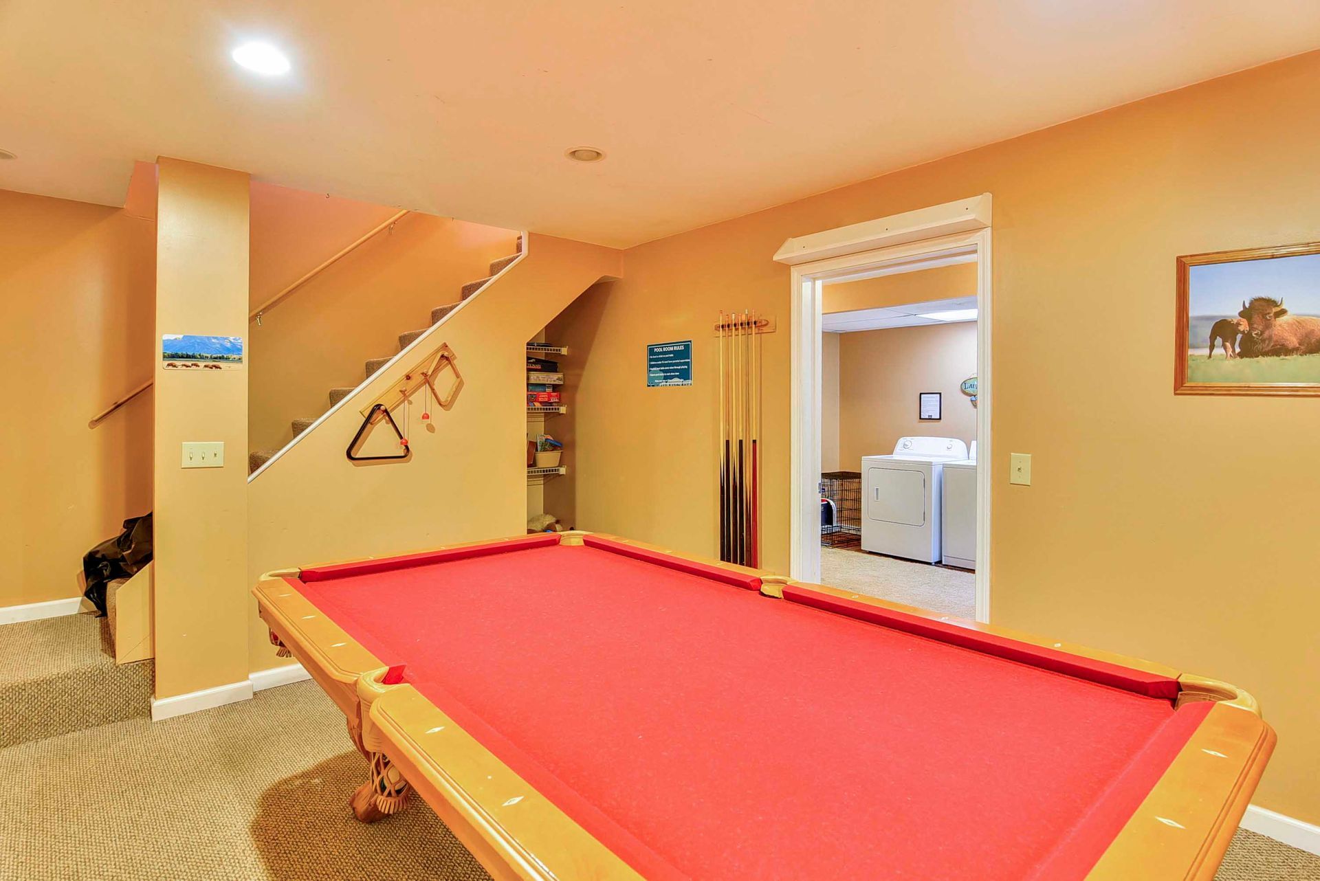 Pool table in a tan-walled basement with stairs. Red felt, cue sticks, doorway to laundry.