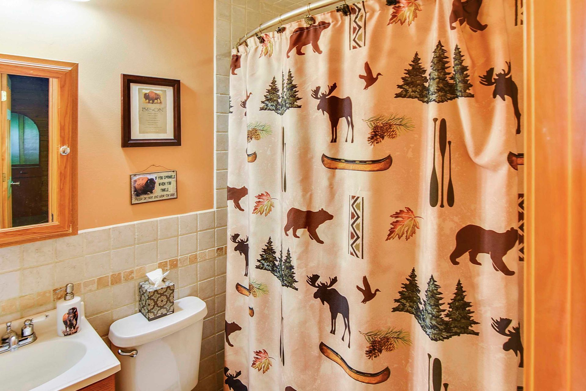 Bathroom with a shower curtain featuring bears, moose, and trees. Orange walls, a toilet, sink, and mirror.