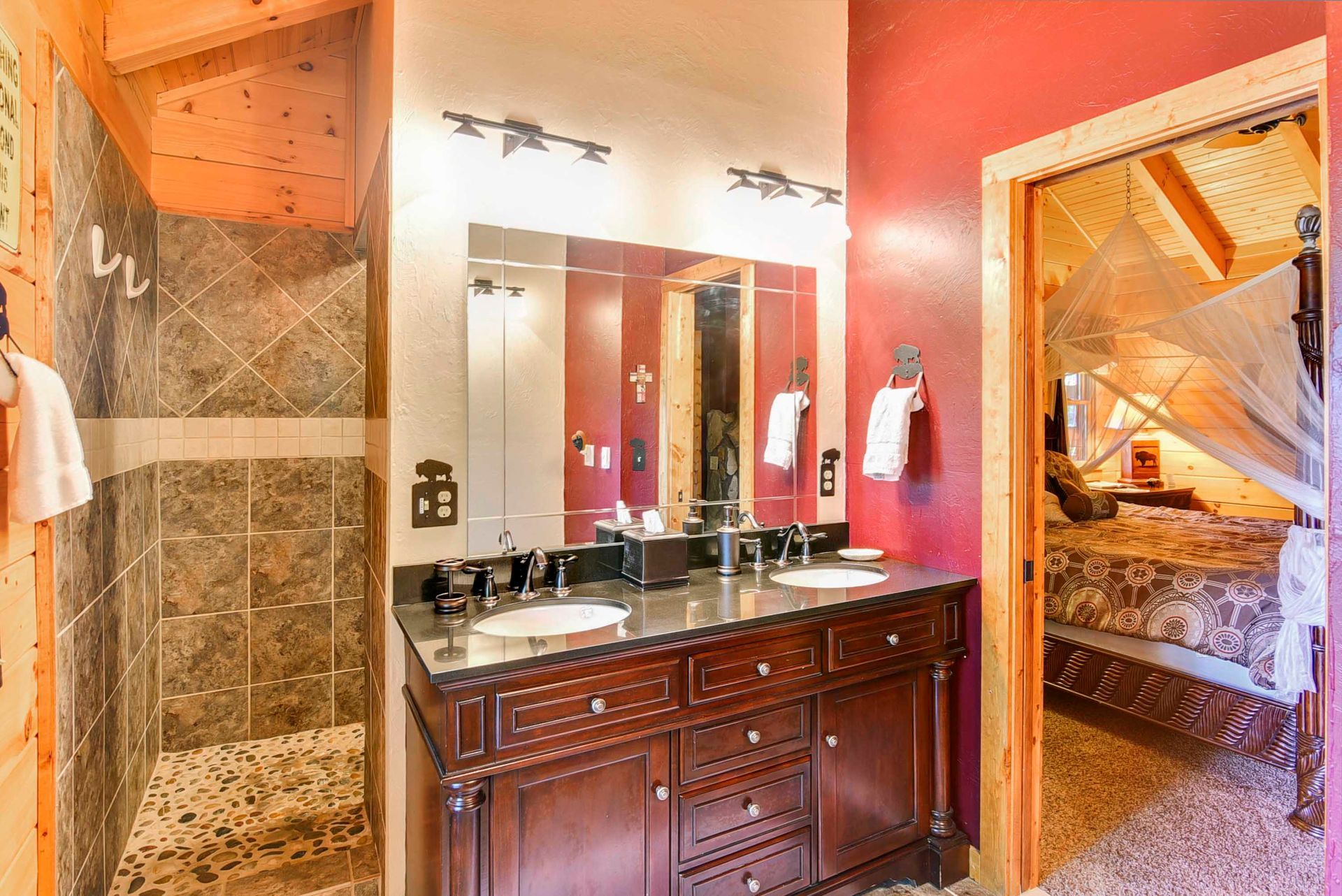 Bathroom with dark vanity, double sinks, red walls, shower stall, and doorway to a bedroom.