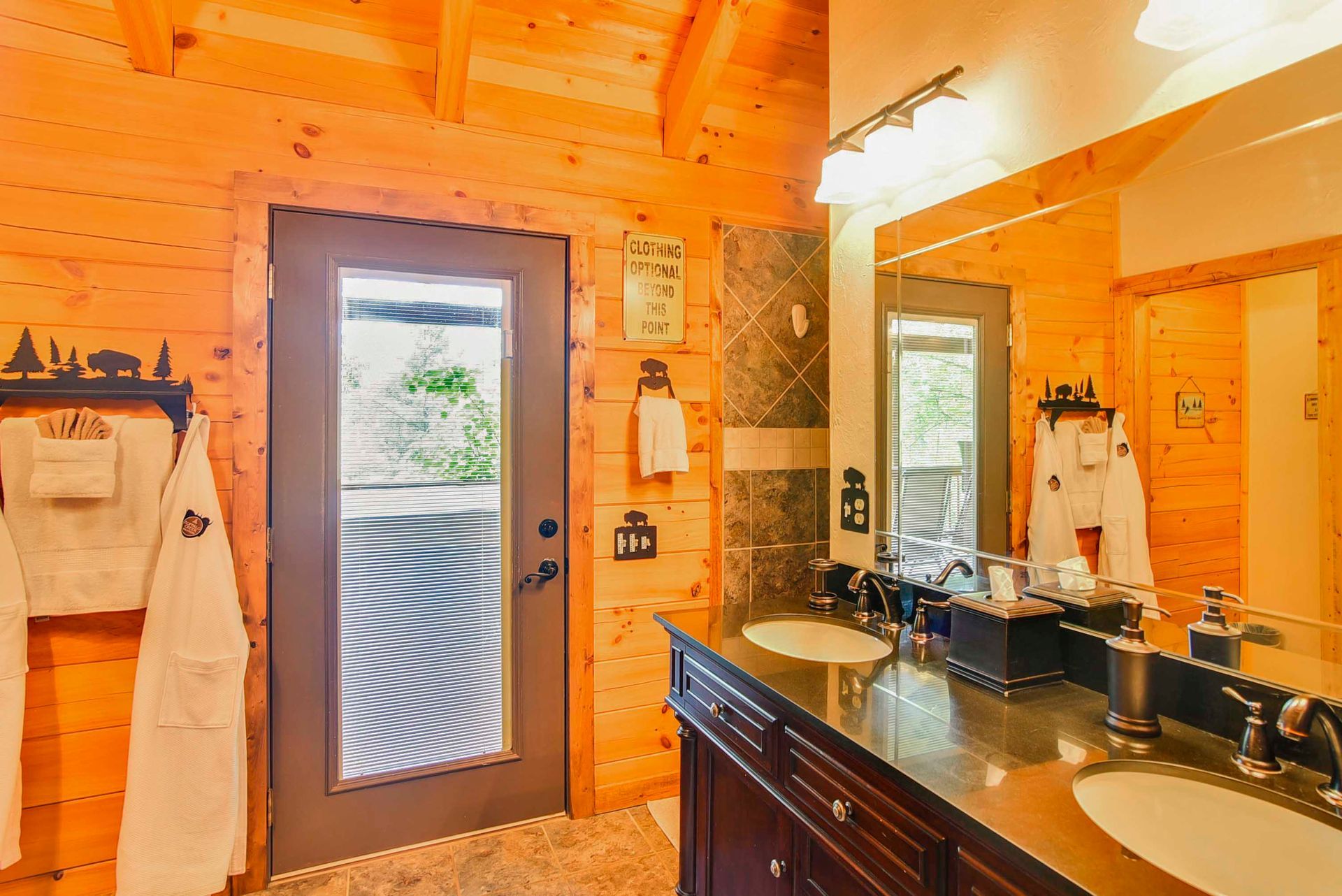 Wooden bathroom with double sinks, a dark brown door, and robes hanging on the wall.