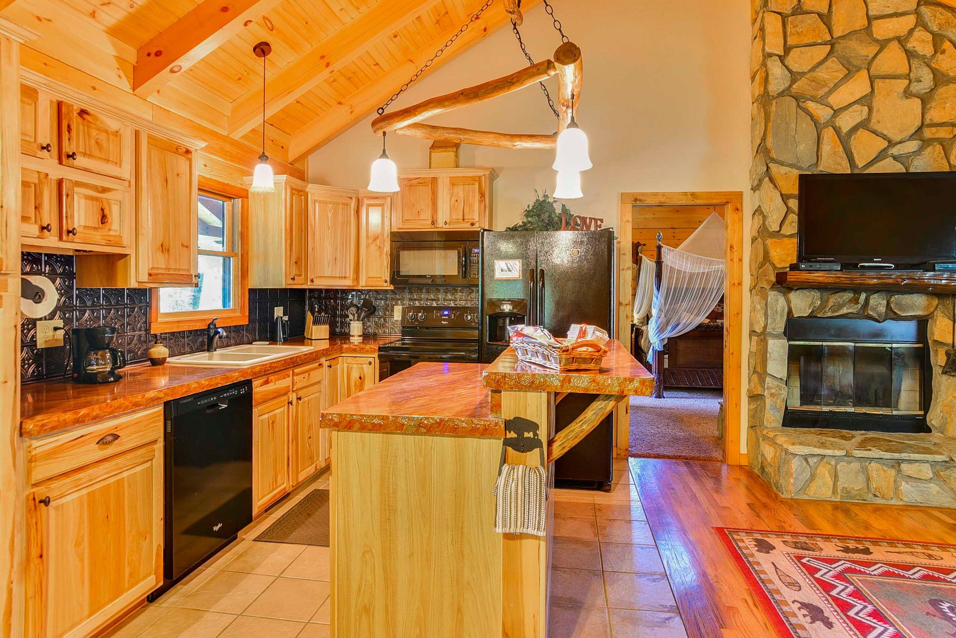 Cozy cabin kitchen with wooden cabinets, island, black appliances, and a stone fireplace.