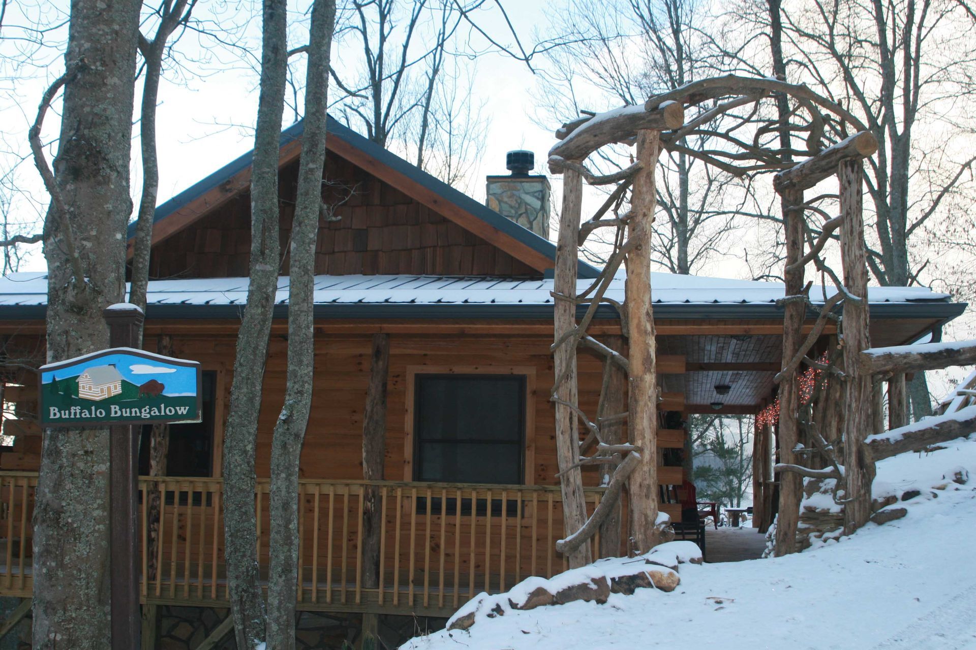 Cabin in the snow with a wooden archway and sign, surrounded by trees.