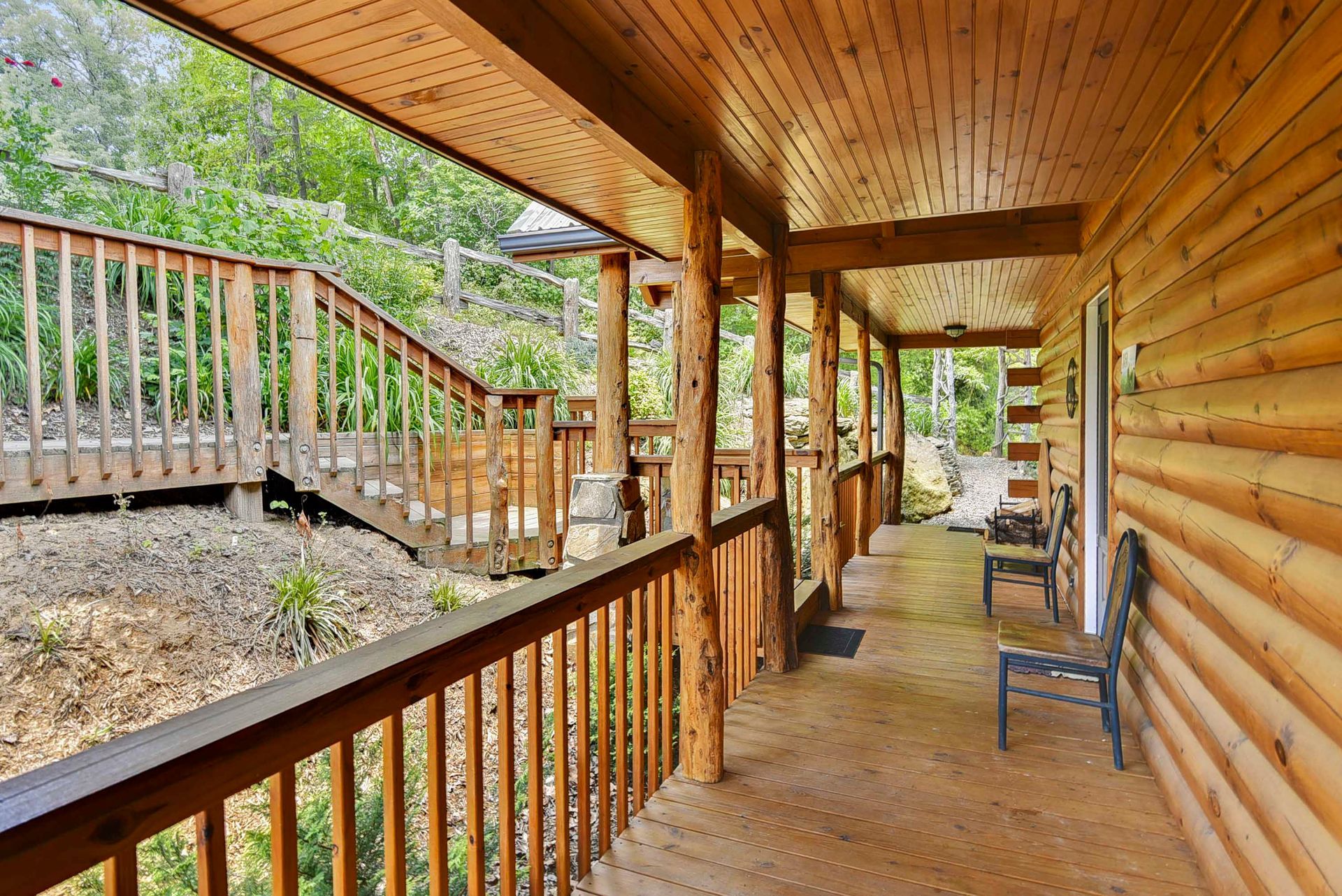 Log cabin porch with wooden railing, steps, and two chairs. Overlooks a hillside.