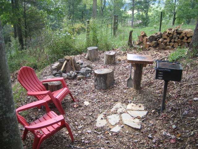 Red chairs around a fire pit with log stumps, a table, and a grill in a wooded area.