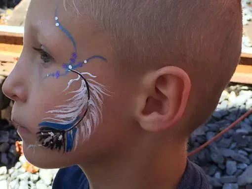 Boy with a feather face paint design on his cheek, blue and white colors, outdoors.