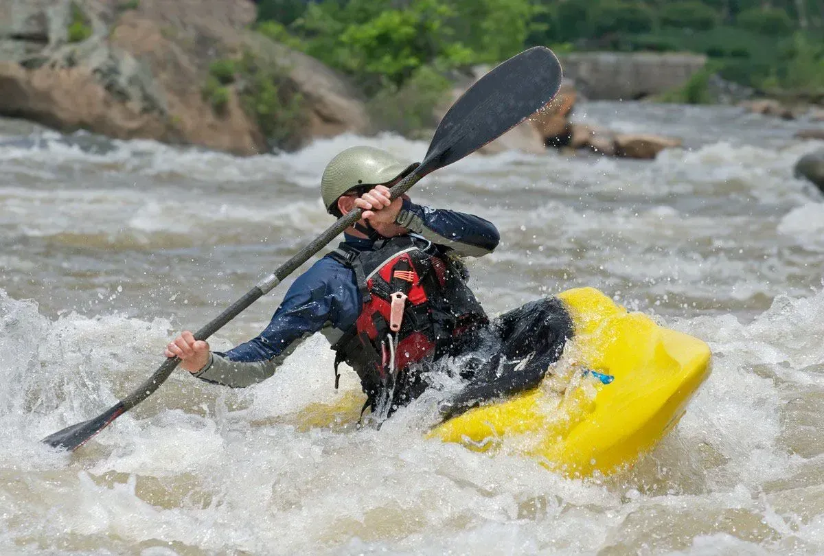 Kayaker in yellow boat paddles through white water rapids; wearing helmet, life vest, and holding a black paddle.